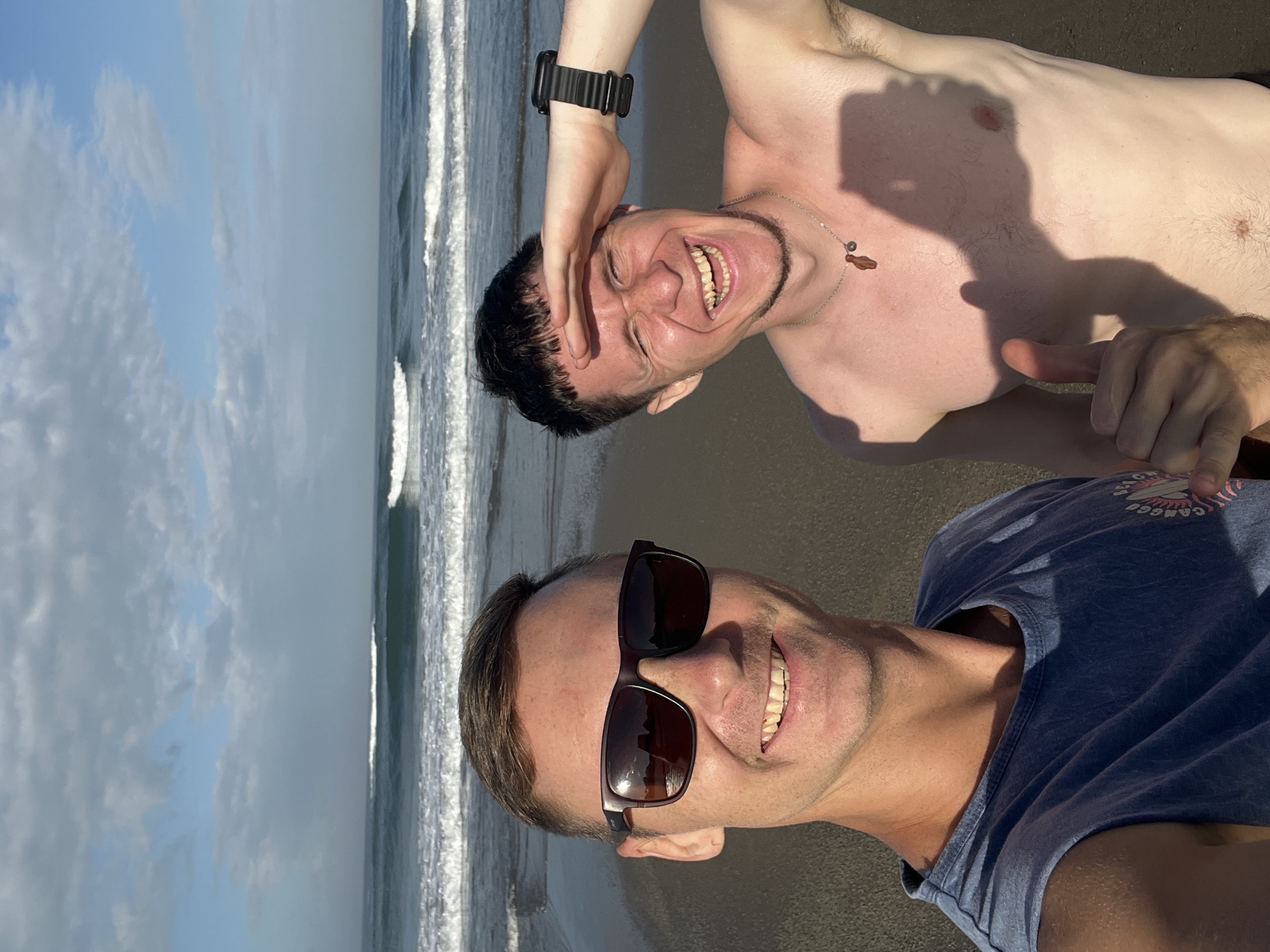 Two men are taking a selfie on a beach in North Kuta, Indonesia. The man on the left is wearing sunglasses and a blue t-shirt, smiling at the camera. The man on the right is shirtless, shielding his eyes from the sun and laughing. The ocean with waves is visible in the background.