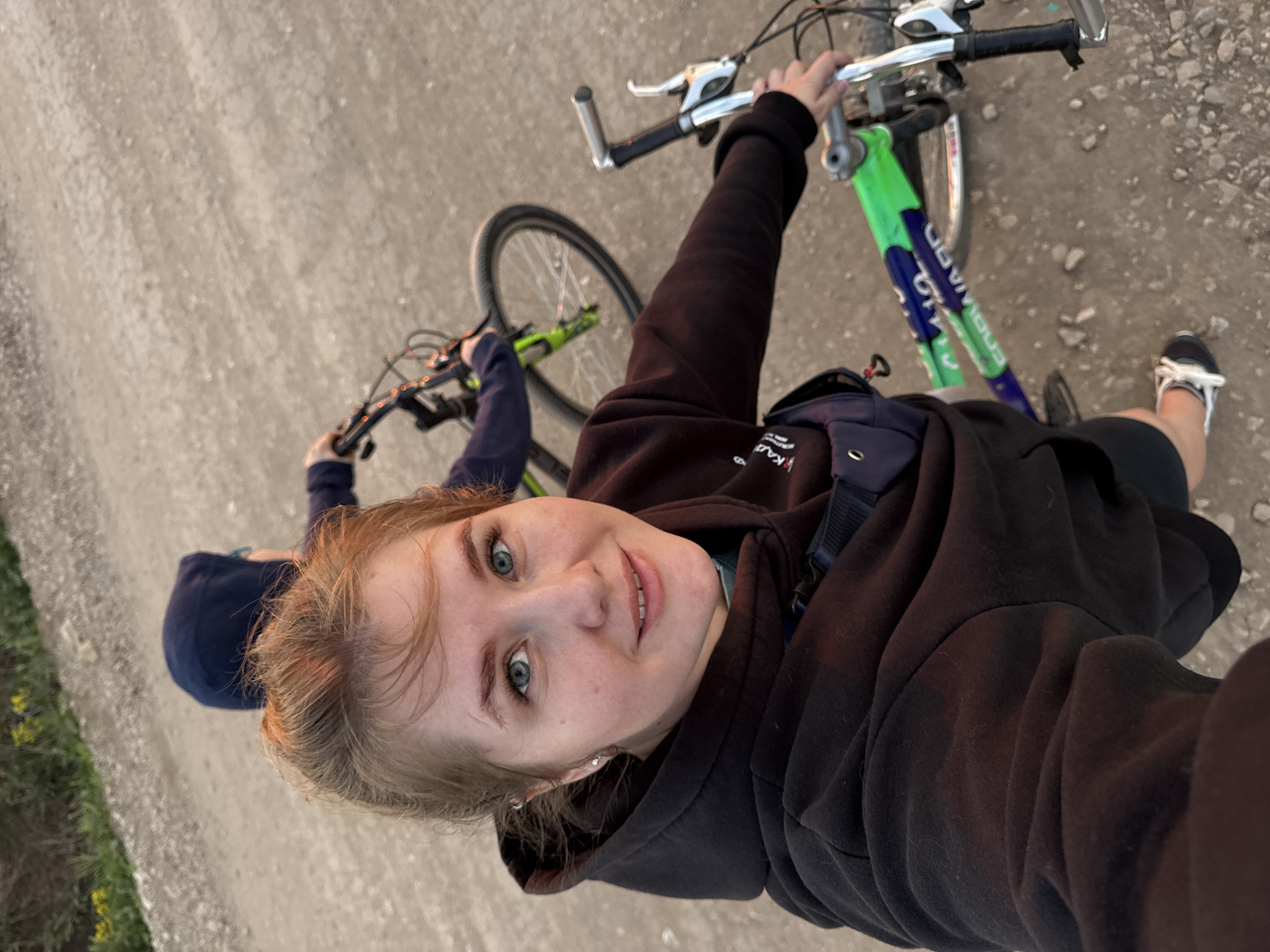 A selfie-style, high-angle shot depicts a young woman and another person riding bicycles on a dirt road. The woman in the foreground has fair skin, light hair, and blue-green eyes. She wears a black hoodie and a shoulder bag, and her bike has a green and blue frame. A second person, partially obscured, rides a similar bike behind her and wears a dark blue hooded sweatshirt. The dirt road, marked by patches of light-colored gravel, extends into the distance. The location appears to be near Горный, Russia due to potential metadata associated with the image, and the environment suggests an outdoor, possibly rural or semi-rural setting. The action involves bike riding.