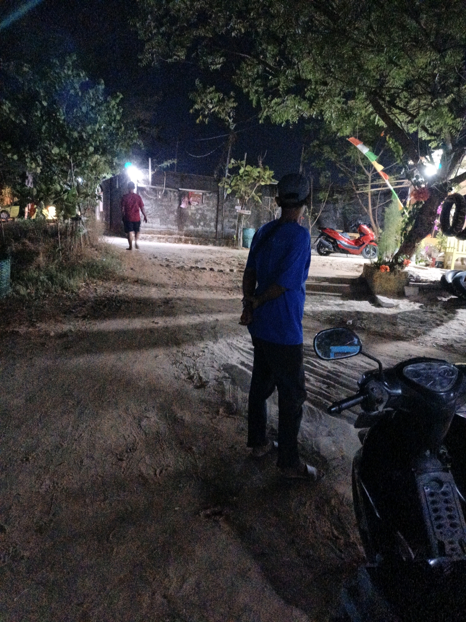 In Cikal, Indonesia, at night, a person stands in the foreground with their back to the camera, wearing a blue long-sleeved shirt and dark pants. They are positioned next to a black motorcycle. In the mid-ground, a person in a red shirt and dark shorts walks away from the camera towards a building with a wall. A bright light illuminates the area. Further in the background, a red scooter is parked. The environment appears to be a sandy or dirt path with trees and foliage surrounding it. A colorful flag or banner is visible hanging from a tree branch. The overall scene suggests a casual outdoor setting at night.