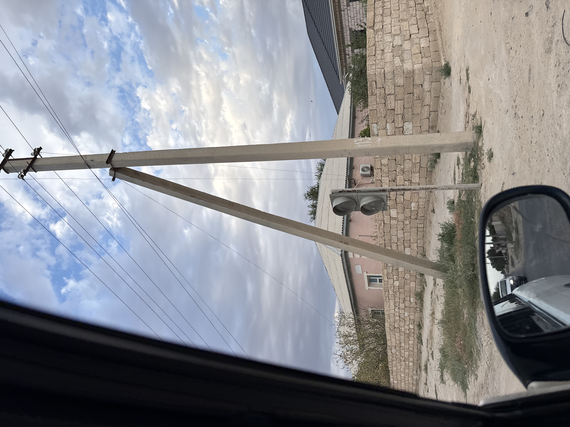 The image shows a street scene from inside a vehicle, looking out at a cloudy sky, a concrete utility pole with power lines, a traffic light, a light pink house with a white roof, and a fence made of light-colored bricks. The ground is unpaved and dusty, with sparse vegetation. A side-view mirror reflects a white car driving on the road behind. The location is identified as Рахат ауылдық әкімдігі, Kazakhstan. There are no people clearly visible in the frame, and no specific actions or interactions are depicted beyond the implied movement of the vehicle. The environment appears to be a rural or semi-urban area.