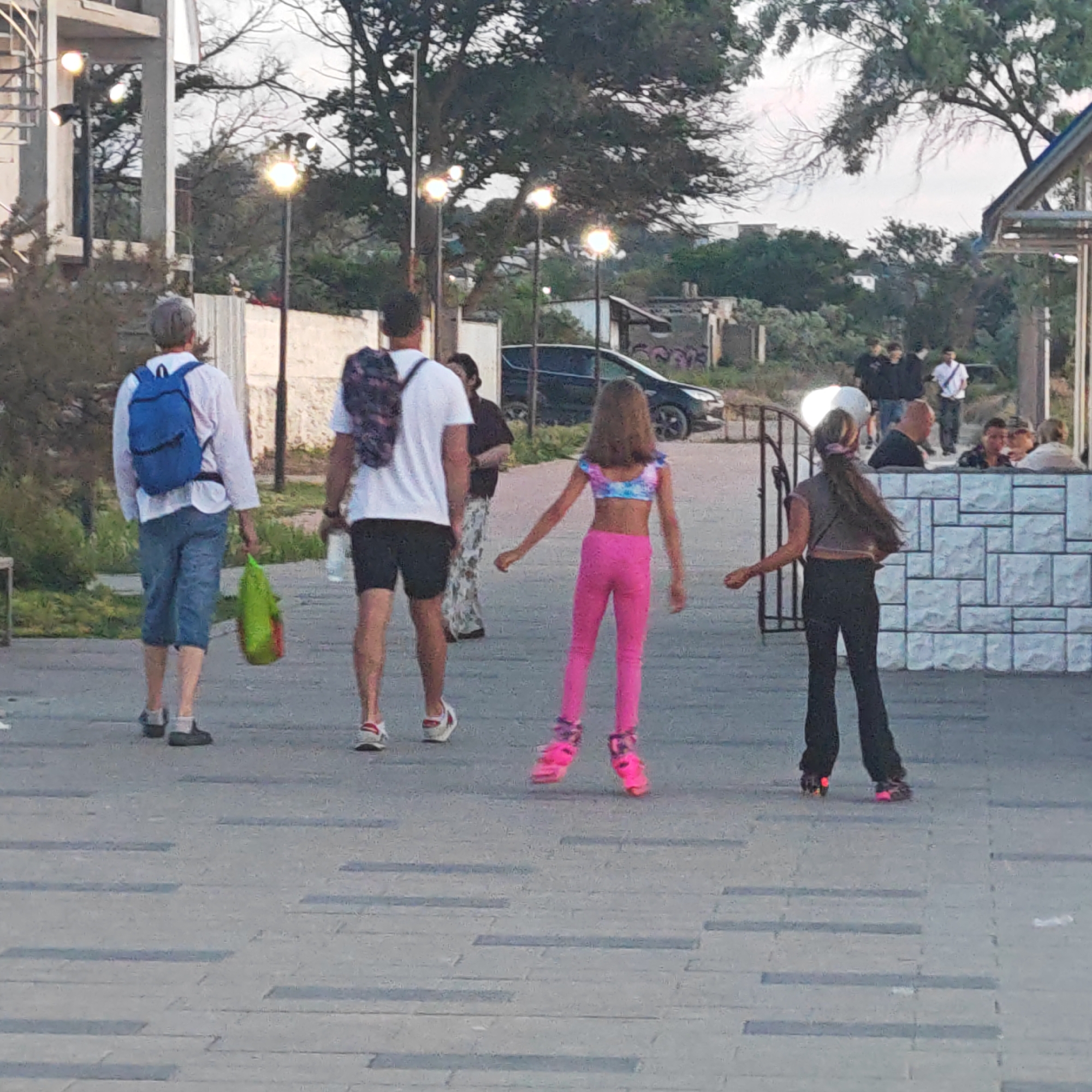 The image depicts an outdoor pedestrian area in городское поселение Щелкино, Ukraine, captured at dusk or early evening. The scene is illuminated by both ambient light and multiple active streetlights.
In the foreground, a paved pathway with alternating light and dark grey rectangular slabs extends into the distance. Two individuals are walking away from the camera. The person on the left wears a white long-sleeved shirt, blue backpack, blue mid-calf trousers, dark shoes with white socks, and carries a bright green bag with orange contents in their left hand. To their right, a second person wears a white short-sleeved t-shirt, dark shorts, white sneakers with white socks, and a patterned backpack.
Directly behind these two, two young girls are moving forward on roller skates. The girl on the left has long brown hair, wears a multi-colored (pink, blue, purple) crop top, bright pink leggings, and pink roller skates with glowing wheels. The girl on the right has long dark hair tied in a high ponytail, wears a dark crop top, dark flared trousers, and dark roller skates with glowing red/orange wheels.
In the middle ground, a dark-colored vehicle (possibly an SUV) is visible on a road behind a low white wall. To the right, a white stone wall with a block pattern separates the pedestrian area from an outdoor seating area where several adults are seated at tables. A dark metal gate is partially visible to the left of this wall.
The background features numerous trees with varying foliage density, some appearing bare-branched. Multiple streetlights with bright, diffused light sources are scattered throughout. A multi-story building is partially visible on the far left. On the right side of the background, a dilapidated structure with visible graffiti ("GOPS") stands near additional distant buildings and vegetation. The sky is a light, overcast grey.