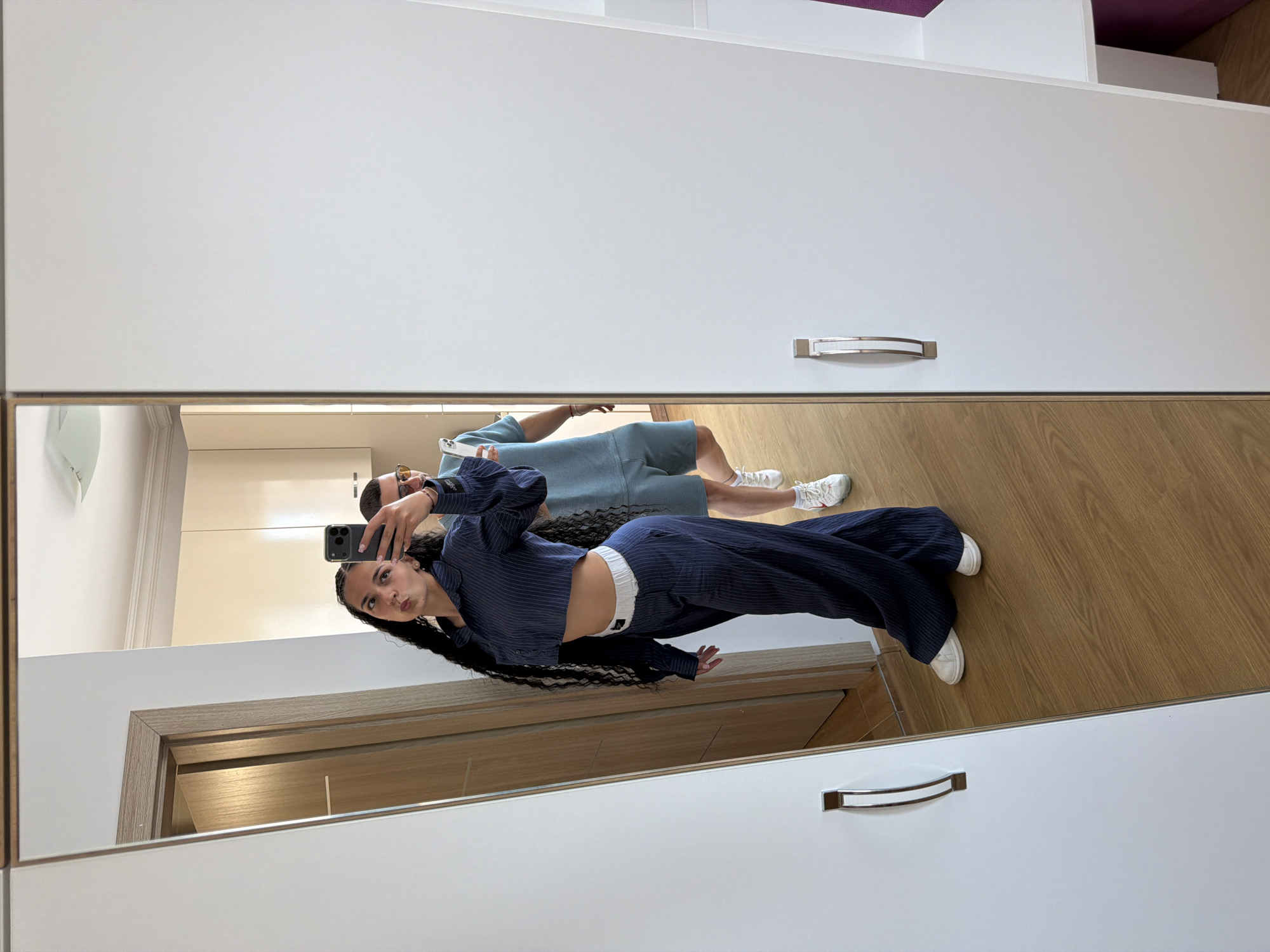 A woman with long, dark, braided hair is taking a mirror selfie in a room. She is wearing a navy blue pinstripe crop top and matching wide-leg pants with a white waistband. A man wearing a light blue t-shirt and shorts is visible behind her in the reflection. The room has light-colored walls and cabinets, and a wooden floor. A white wardrobe with a silver handle is to the right of the mirror. The woman is posing with her phone in her hand, and her mouth is puckered. The man in the reflection is wearing sunglasses. The image appears to have been taken in a residential setting, possibly an apartment or house, in Nesebar, Bulgaria, as indicated by the surrounding context.