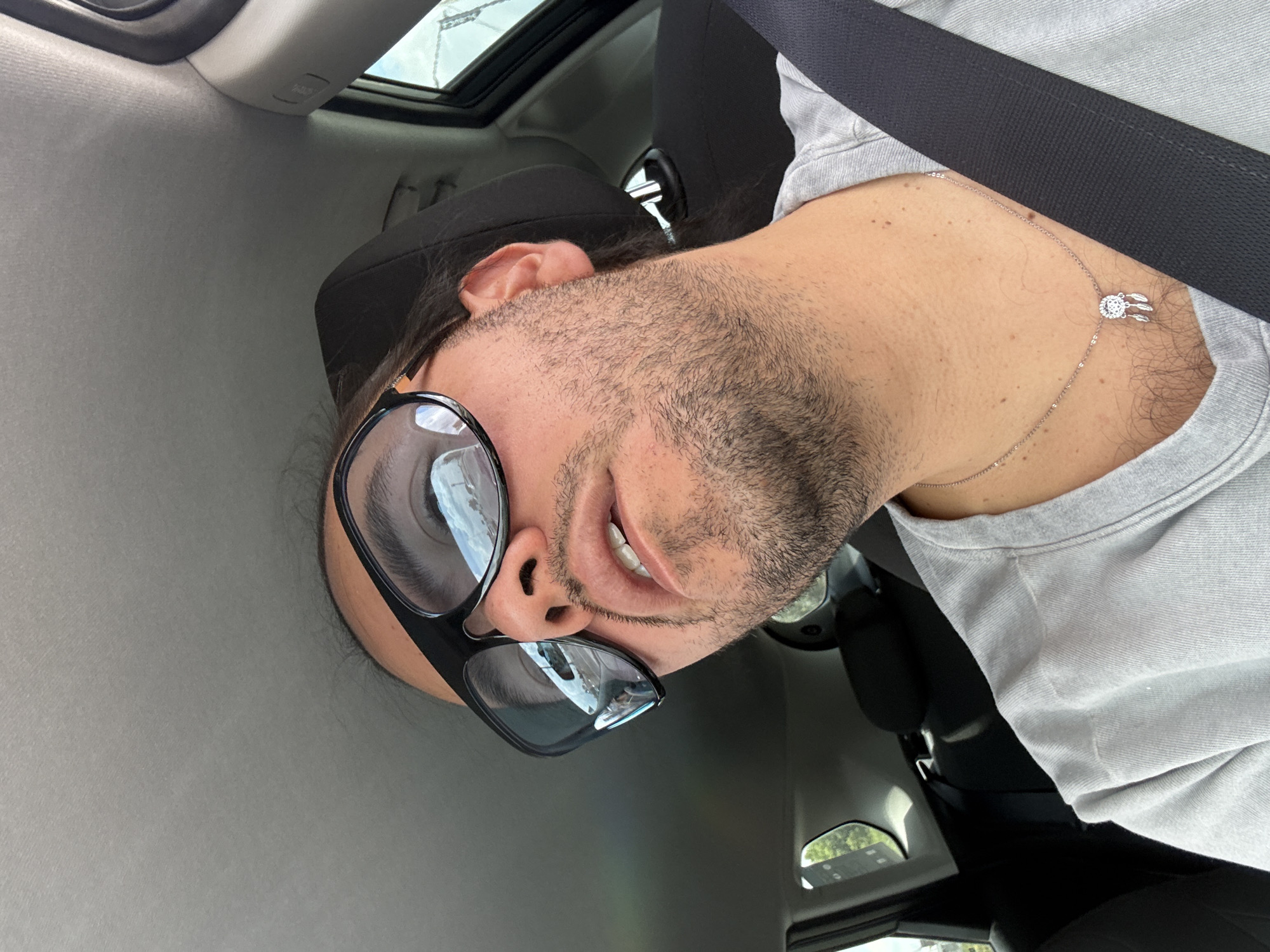 A selfie taken from a low angle inside a vehicle, potentially a car. The subject is a light-skinned male wearing a grey shirt and a black seatbelt. He has dark hair and a beard. He's wearing large black sunglasses and a silver necklace with a dreamcatcher pendant. The interior of the car is visible, including part of the sunroof. The location within Barrio de Nuevo León, Mexico, is not discernible from the image.