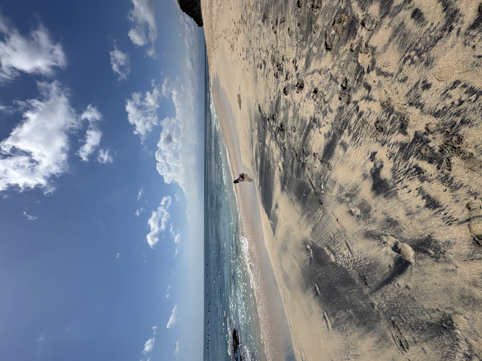 The image shows a beach with alternating bands of light and dark sand in Kuta Selatan, Indonesia. The water is clear and blue with small waves. A single person is visible at the water's edge. The sky is blue with scattered white clouds. The angle of the photograph is unusual with the horizon appearing vertical.