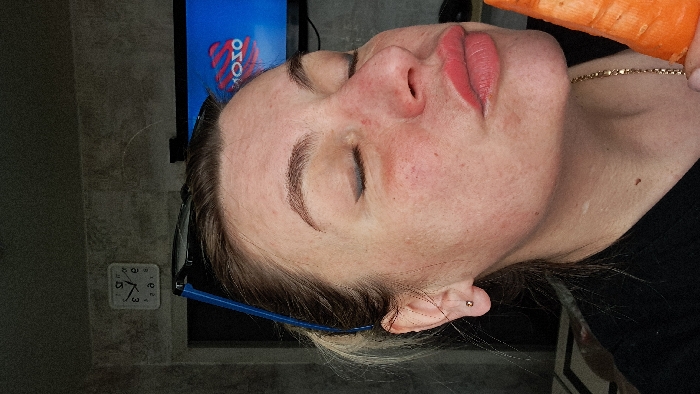 The scene is an indoor domestic setting in Bel'kovo, Russia. A female subject, of apparent adult age, is captured in a mid-shot, her head tilted slightly, with her face in profile, directed towards the lower right. Her eyes are closed, with visible black winged eyeliner. Her lips are pursed and colored with a light red lipstick, positioned in close proximity to a large, orange, raw carrot held upright in her right hand, which is mostly out of frame. She exhibits fair skin with visible pores, minor redness on the cheeks, and well-defined eyebrows. Blue-framed glasses are resting on top of her head, and a small gold-colored stud earring is visible in her left earlobe. A thin gold-colored chain is worn around her neck, partially visible over a dark, indeterminate garment. The background consists of a dark, textured wall. To the upper left, a white square wall clock with black hands displays a time approximating 11:45 or 12:45. To the upper right, a television screen is visible, displaying a blue background with a white and red graphic, possibly a logo, which appears as "MOTO" or "OZO" in reverse.
