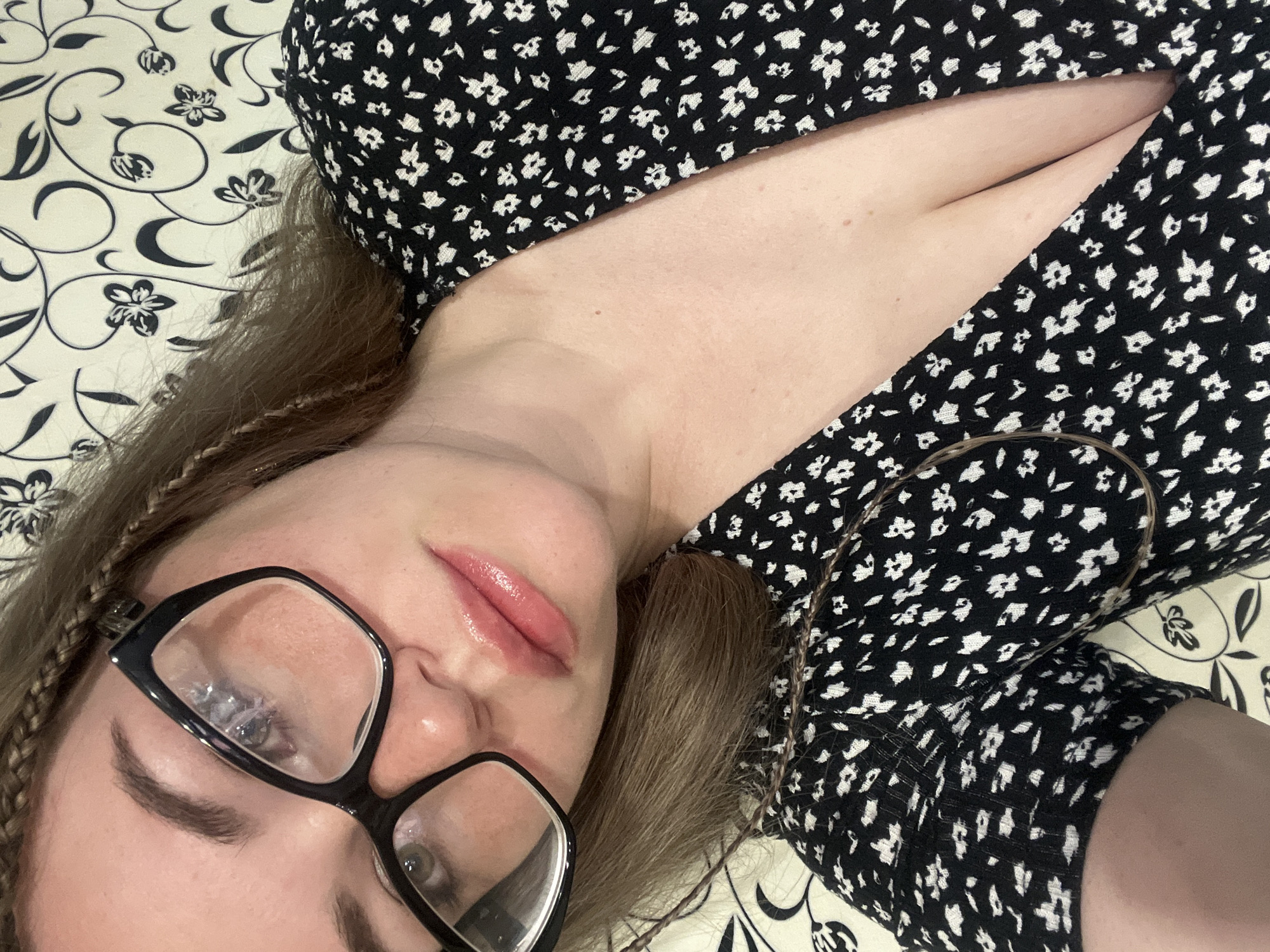 A woman with light skin and brown hair is lying down. She is wearing black-framed glasses and a black and white floral patterned dress. The background is a cream-colored fabric with a black floral pattern. There is no information to suggest that the image was taken in the city of Chaikovsky, Russia.