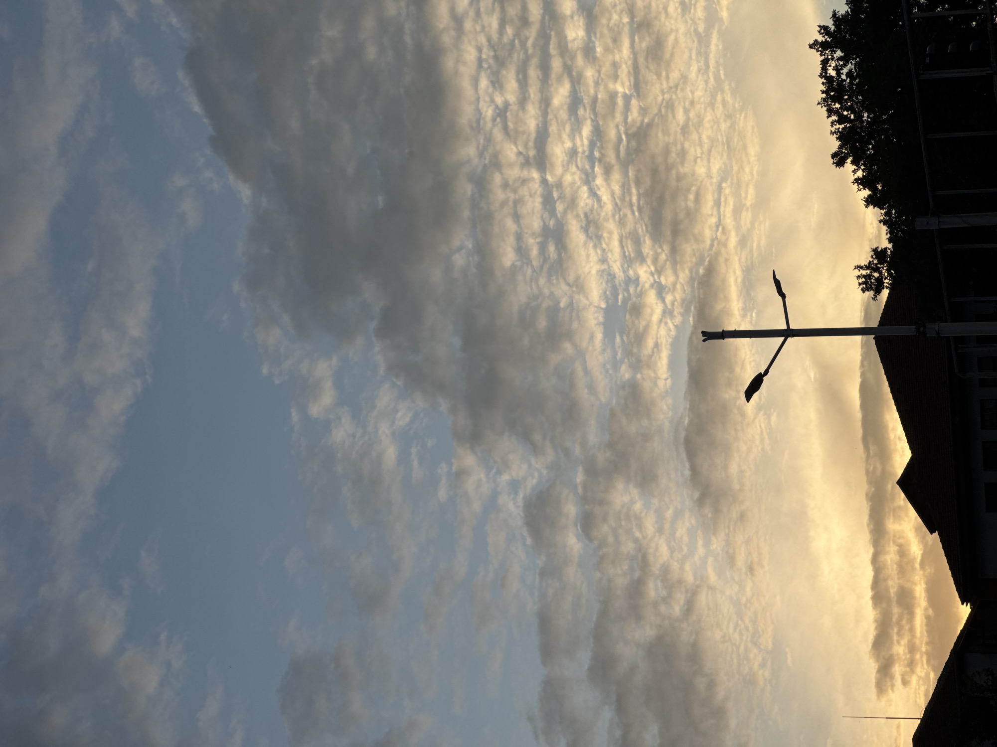 The image depicts a sky filled with clouds, illuminated by the warm light of sunset. The clouds are varied, with some appearing wispy and others more dense, catching the golden hues of the setting sun. In the foreground, silhouetted against the bright sky, is a street lamp with a distinctive crossbar. To the right, the dark silhouettes of trees and the roofline of a building are visible, suggesting an urban or suburban setting. The overall scene evokes a sense of calm and the end of a day. The time of day is clearly sunset due to the lighting and colors of the sky. The weather appears clear apart from the clouds. There is no visible text in the image.