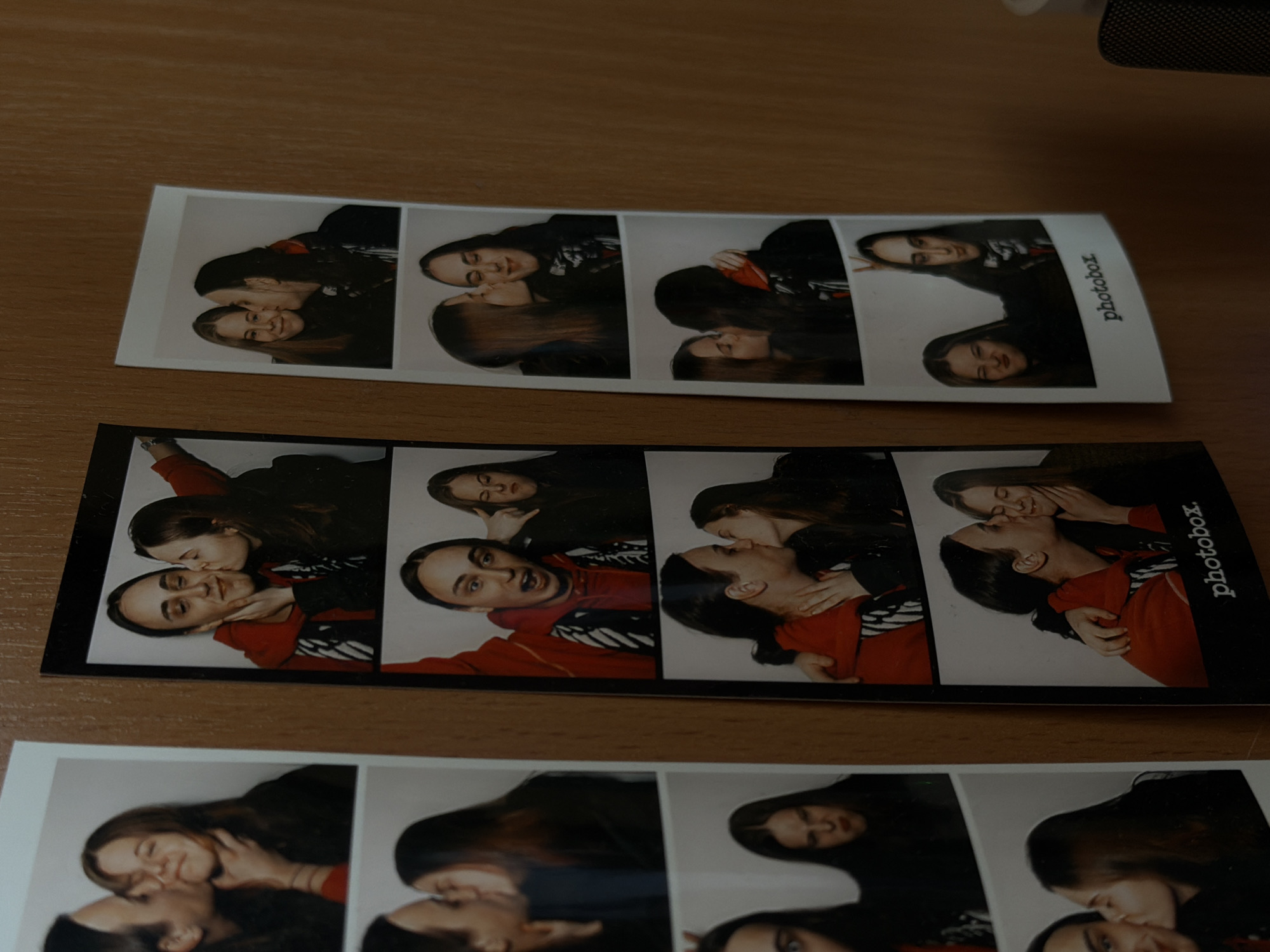 The image shows three strips of photobooth pictures laying on a wooden surface. The photobooth strips depict two individuals, presumably a couple, interacting in various poses. Some frames show them kissing or embracing. In one frame, one individual makes bunny ears behind the other's head. The strips are labeled "photobox" along the right edge. The location of the image, Shul'gino, Russia, is not depicted in the picture.