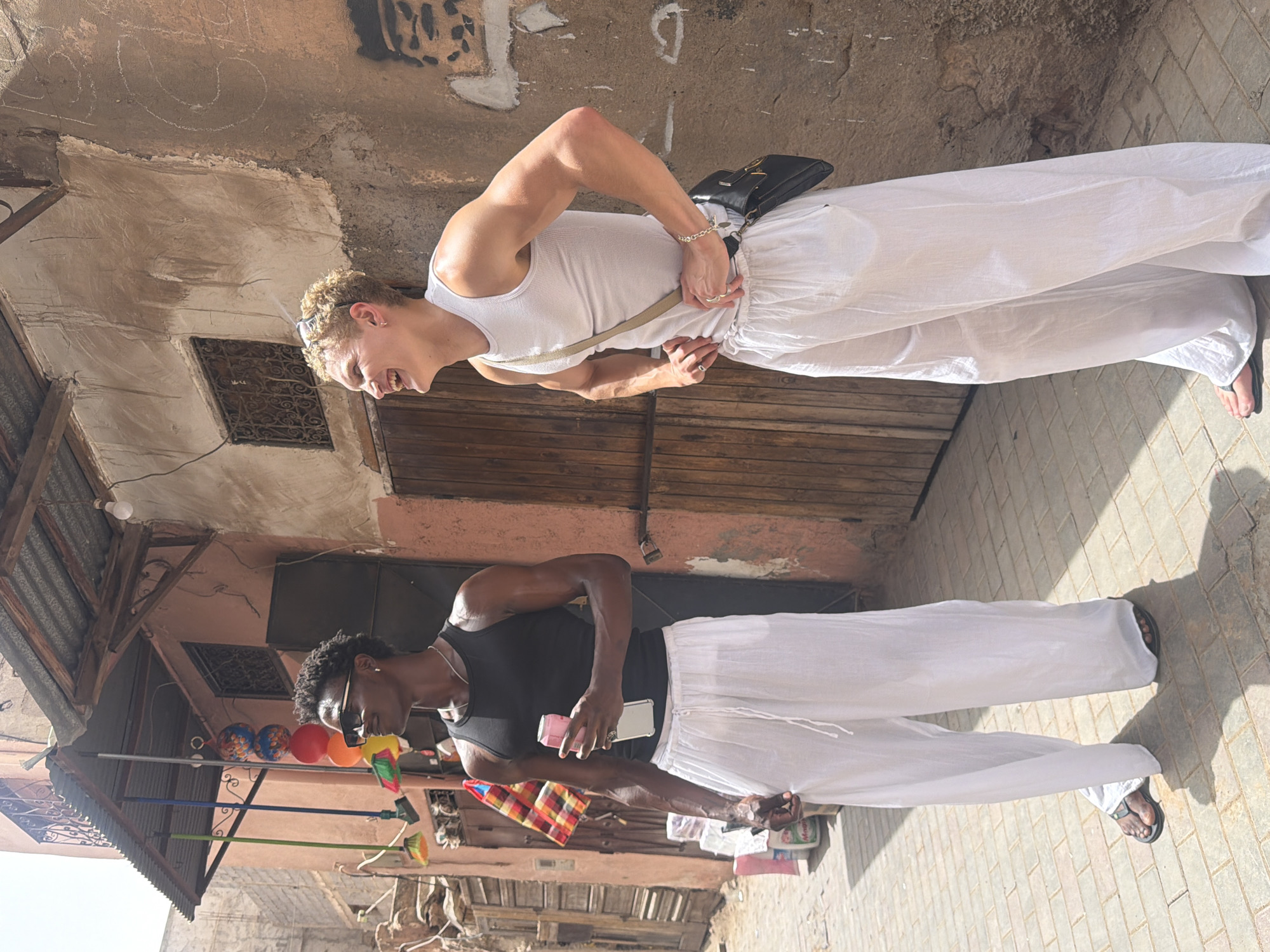 Two young men are standing on a paved street in a historic Moroccan city, likely Marrakech, given the architecture. The man on the left has dark skin, short curly black hair, and is wearing sunglasses, a black tank top, and loose white pants. He is holding a pink can and a phone. The man on the right has light skin, short curly blonde hair, and is wearing a white tank top, loose white pants, and sunglasses on his head. He has a black bag slung across his chest. The background shows a weathered building with wooden doors and windows, a corrugated metal awning, and some hanging decorations. The sunlight casts strong shadows on the ground. The scene depicts a casual moment between two individuals in a vibrant, sun-drenched urban environment.