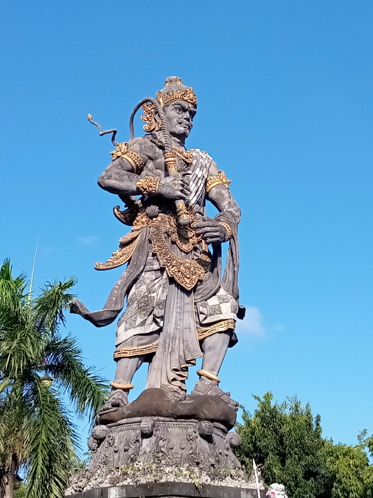 A low-angle, full-shot photograph shows a large stone statue of a Balinese deity or warrior, predominantly gray with gold accents, standing against a clear, vibrant blue sky. The statue, elaborately adorned with intricate carvings and golden decorative elements, holds a weapon in its hands. The figure wears a headdress and ornate clothing, with a rope coiled over its shoulder. The statue stands on a detailed stone pedestal adorned with decorative carvings. To the left of the statue, a palm tree with lush green fronds is partially visible, and to the right, a dense green canopy of trees forms the background. The overall impression is one of grandeur and cultural significance, likely located in Blahbatuh, Indonesia, given the prominent Balinese artistic style.
