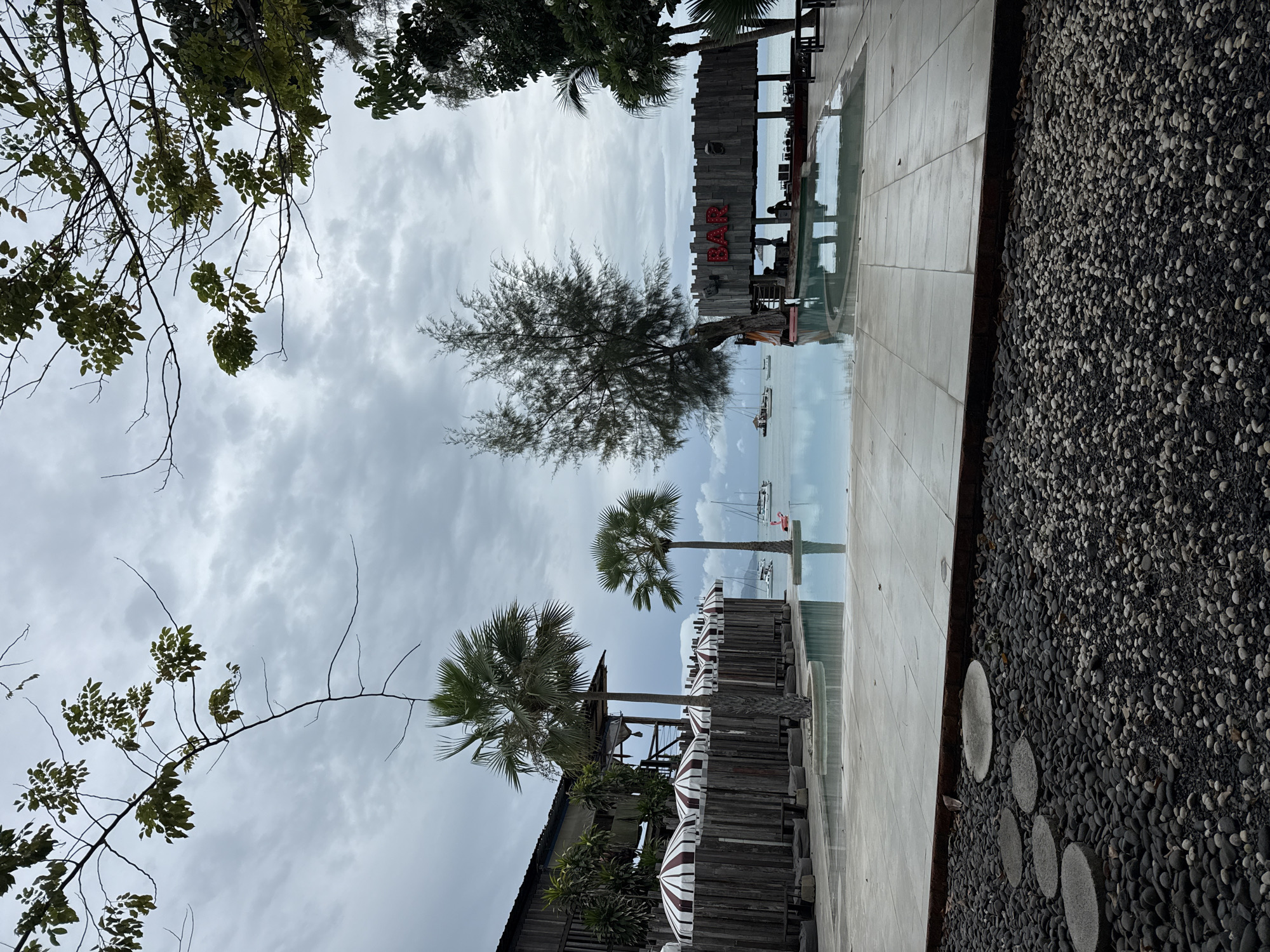 The image shows an outdoor bar and swimming pool area, likely in Ko Samui District, Thailand, under a cloudy sky. The foreground features a pathway of circular concrete stepping stones laid into a bed of dark and light-colored pebbles. Beyond this is a tiled patio area bordering a large, tranquil swimming pool with clear, reflective water.
To the left, a rustic wooden building with a covered area and striped umbrellas is visible. Several lounge-like seating areas with beanbag chairs are arranged along a wooden fence. Palm trees with green fronds are scattered throughout the scene, two prominently in front of the building.
In the background, the ocean stretches out with several sailboats and what appears to be a pink inflatable object floating in the water. A prominent wooden bar structure stands to the right of the pool, with the word "BAR" lit in red neon letters above it. Silhouettes of people can be seen inside the bar area. A tall, thin tree with sparse foliage stands next to the bar. The overall atmosphere suggests a relaxed resort or beachside establishment.