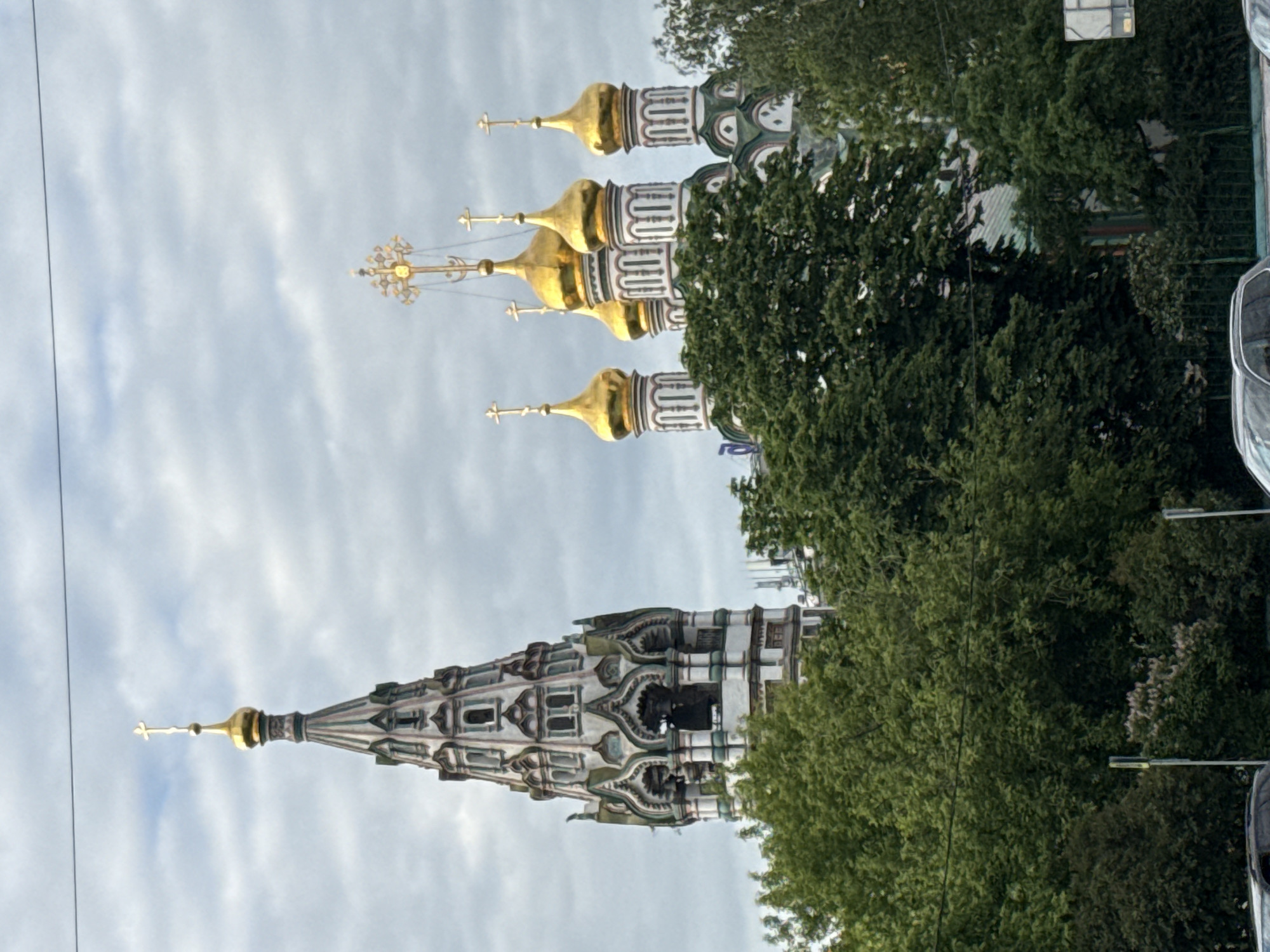 The image captures a vibrant architectural landscape featuring ornate Russian Orthodox church structures, framed by abundant green foliage, under a cloudy sky.
In the upper right portion of the frame, a cluster of at least four, possibly more, gleaming golden, onion-shaped domes topped with crosses stands out. These domes crown multi-tiered structures adorned with intricate white and green patterned facades, characteristic of traditional Russian ecclesiastical architecture. To the upper left, a tall, slender spire rises prominently. It features a distinctive, ornate pattern of white, grey, and possibly green geometric motifs, culminating in a golden finial and cross at its apex. This spire appears to be part of a larger building, mostly obscured by trees.
Dense, lush green trees dominate the lower and central sections of the photograph, partially obscuring the lower parts of the religious buildings and softening the architectural lines. The sky, occupying the upper background, is overcast with soft, diffused clouds, indicating a daytime shot, likely late morning or afternoon. A thin black line, possibly a power cable, stretches across the sky on the far left. Through gaps in the foliage on the far right and bottom, indistinct elements that might be other structures or vehicles are faintly visible.
The architectural styles, particularly the iconic onion domes and detailed spires, strongly suggest the location is Moscow, Russia, consistent with the provided context. The scene conveys a sense of historical grandeur, cultural richness, and the integration of majestic architecture with nature. No visible text is present in the image.