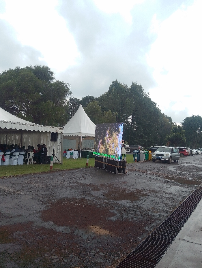 This image captures an outdoor event taking place in Gitaru, Kenya, under an overcast, cloudy sky. The ground is wet and appears to be a mix of gravel and mud with puddles, suggesting recent rainfall.
On the left side of the frame, a large white rectangular marquee is erected, under which numerous people are seated on rows of white chairs, some decorated with red sashes. They appear to be attending a gathering or presentation. A large black speaker on a tripod stand is positioned in front of this tent. Adjacent to the main marquee, a smaller, pointed white canopy tent stands alone.
Centrally located is a prominent, large, vertical LED screen displaying what looks like a vibrant natural scene, possibly trees or foliage. The screen is mounted on a dark, multi-layered base, which might house associated equipment.
To the right of the LED screen, several cars are parked on the wet, gravelly surface, including a silver SUV. Near the parked cars, a cluster of colorful waste bins (green, blue, yellow) is visible. A long, dark metal drainage grate extends along the bottom right edge of the frame. Small red and white striped poles are intermittently placed on the ground, possibly marking boundaries or pathways.
The scene depicts an organized outdoor event, equipped with temporary structures for shelter and seating, as well as a large screen for visual presentations. The damp conditions are a notable aspect of the setting. No discernible text is visible in the image.