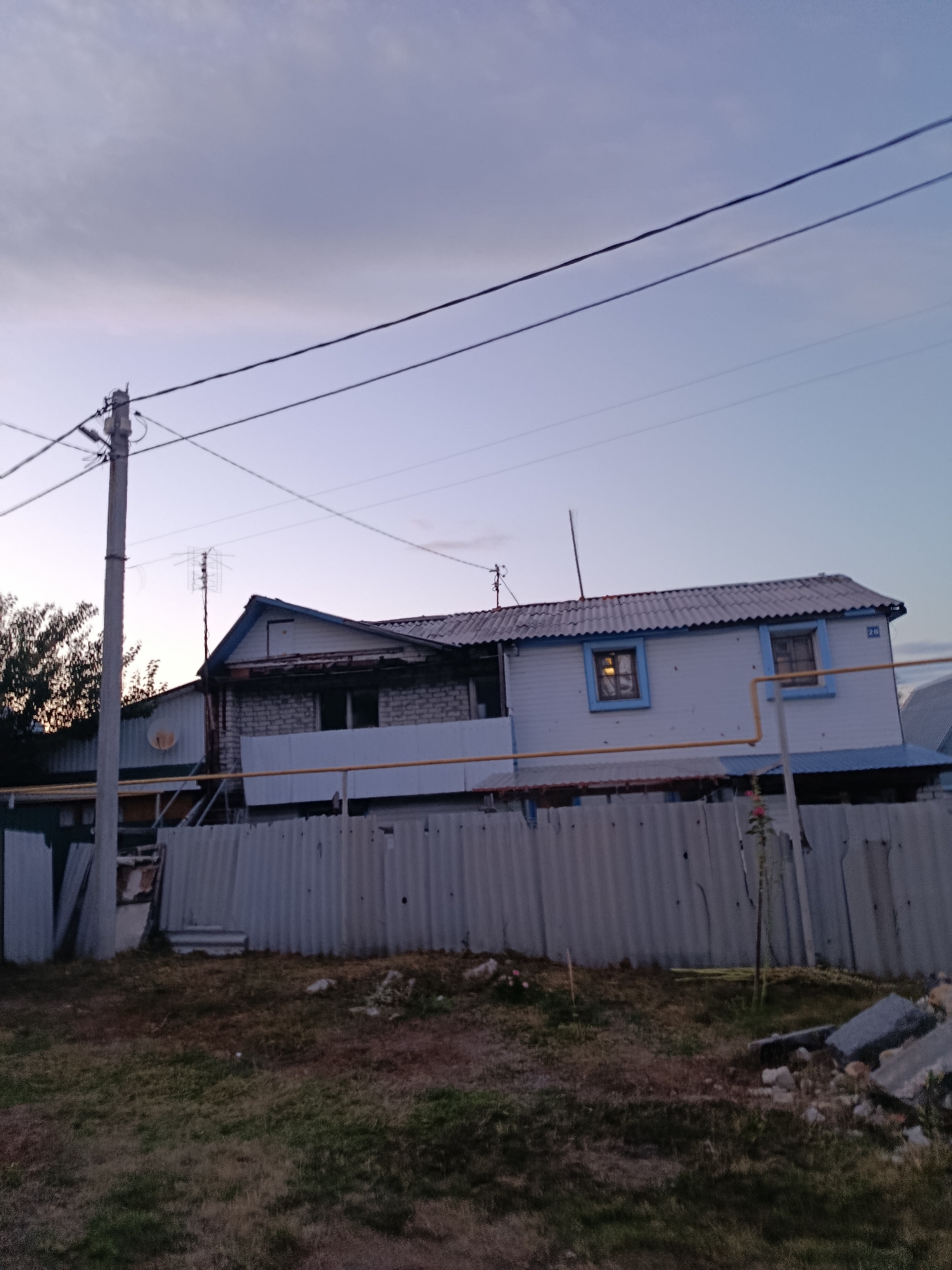 The image shows a residential house with a corrugated metal fence in front of it, in Saratov, Russia. The house is a two-story structure with a light blue trim around the windows. The sky is overcast with a light purple hue, suggesting it is either dawn or dusk. There are power lines running across the sky, and a utility pole on the left side of the image. The fence is made of corrugated metal sheets and appears to be somewhat damaged. There is grass and some debris in the foreground, indicating a rural or semi-urban setting. The overall scene depicts a modest dwelling in a somewhat unkempt environment.