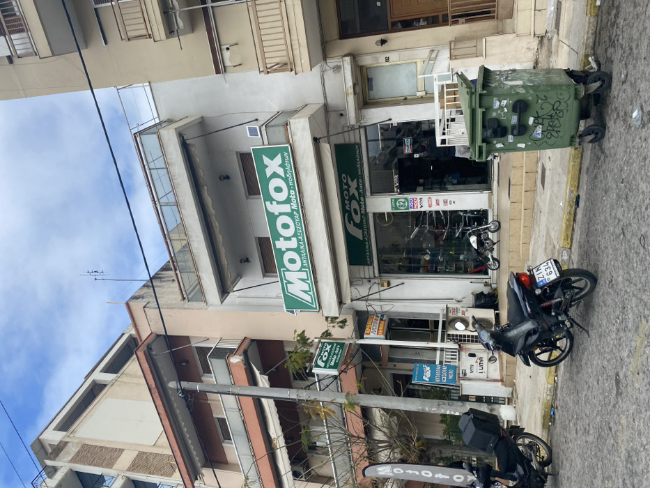 The image depicts a daytime street scene in Nikea, Greece, centered on a commercial establishment named "Motofox." The setting is an urban environment with multi-story buildings, characterized by balconies and typical Mediterranean architecture. The sky is partly cloudy.
The primary subject is the "Motofox" store, which occupies the ground floor of a building. Large green signs prominently display the store's name, "Motofox," in a distinctive font. One main sign reads "Motofox ΑΝΤΑΛΛΙΚΑ-ΑΞΕΣΟΥΑΡ Moto - ποδηλάτων," indicating it sells "Spare Parts-Accessories Moto - Bicycles." A smaller sign below it further specifies "ΑΝΤΑΛΛΙΚΑ ΑΞΕΣΟΥΑΡ Moto-Auto - ποδηλάτων," including "Auto" (cars) in its offerings. The shop window displays various motorcycle accessories and parts.
On the street and sidewalk in front of the store, two motorcycles are parked. One black motorcycle, visible in the foreground, has a license plate reading "ZIN 634." Another motorcycle is partially visible behind a street pole, featuring a white banner with the "MOTO FOX" logo. Adjacent to the store entrance, a large green trash bin, covered in stickers and graffiti, is visible. A utility pole with numerous wires stands near the sidewalk. Other visible text includes "VPR," "LUMA," and "LIQUI MOLY" inside the shop window, likely brand names, and the number "82" on one of the buildings. No people are visible, suggesting a quiet moment on the street.