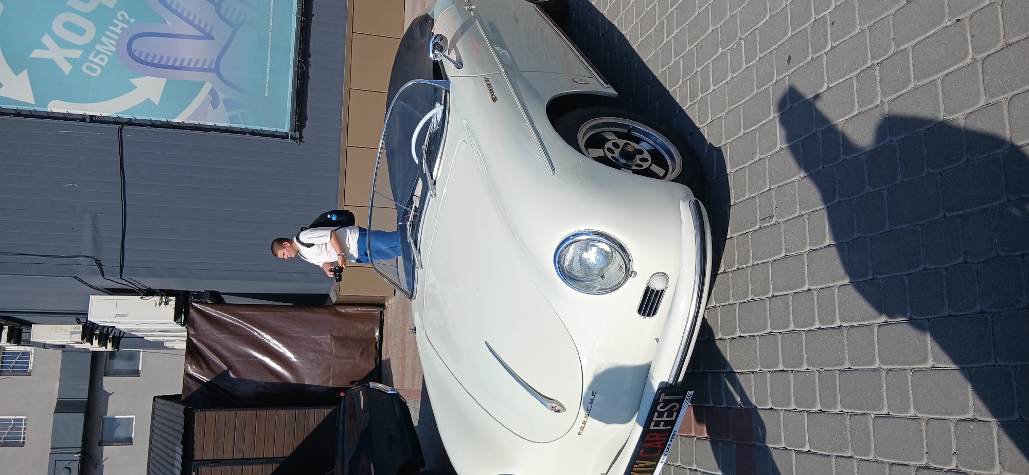 This media file captures a white vintage car, likely a Porsche, parked on a cobblestone surface. The car's sleek design, including its round headlights and chrome accents, is prominently displayed. The image is taken outdoors, under bright sunlight, suggesting a daytime setting with clear weather. A person, wearing a white t-shirt and blue jeans and carrying a backpack, is standing near the car, appearing to take photographs or observe it. The background includes a building with a dark gray facade and a large banner with blue and white graphics. Some greenery and brown tarps are also visible, hinting at an outdoor event or exhibition. A sign at the bottom of the car reads "XIV CAR FEST," indicating that this is part of a car show or festival. The overall mood is one of appreciation for vintage automobiles and community gathering.