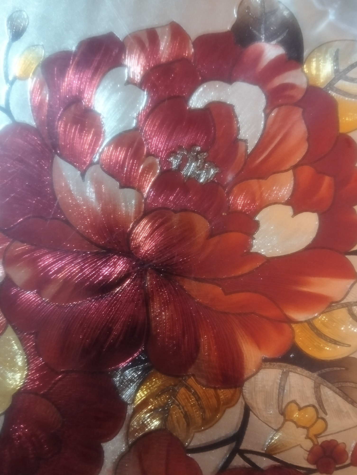 The image is a close-up, overhead shot of a detailed floral embroidery. The embroidery features large, overlapping petals in shades of deep red, crimson, orange, and white, with accents of gold and silver thread. The petals have a shimmering, metallic appearance, suggesting the use of silk or satin threads. The center of the flower appears to have a cluster of small, intricate silver details. The background is a pale, off-white fabric, possibly silk or satin, which also has a subtle sheen. The overall impression is one of luxurious craftsmanship, likely part of a larger textile piece such as a garment, tapestry, or decorative panel. There is no indication of a specific location in Payagaboh, Indonesia, or any environmental context beyond the textile itself.