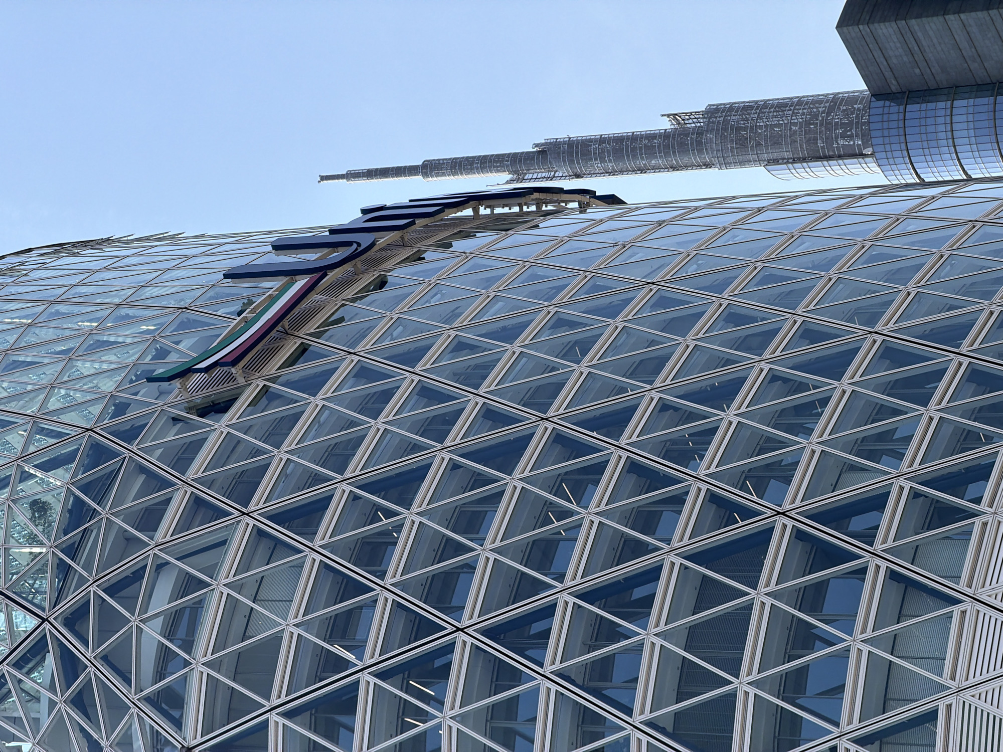 The image shows a close-up view of a modern glass-paneled building with a triangular facade in Milan, Italy. A sign with Arabic lettering and the colors of the UAE flag is visible on the building's exterior. In the background, the iconic Burj Khalifa skyscraper can be seen, suggesting the image may be a mislabeled or intentionally misleading depiction. The sky is clear and blue, indicating a sunny day. The focus is on the architectural details of the building and its signage, with the Burj Khalifa serving as a contrasting element in the distance.
