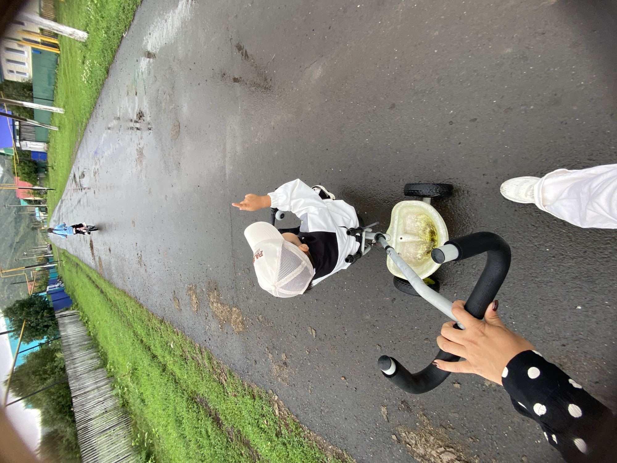 In Maloarslangulovo, Russia, on a wet asphalt road, a young child, wearing a white jacket and a light-colored cap, sits in a white tricycle. The child is facing away from the camera and has their left arm raised with a finger pointing upwards. In the foreground, a person's hand, wearing a black and white polka dot sleeve, is gripping the tricycle's handlebars. To the right of the tricycle, another person's lower legs and white sneakers are visible, suggesting they are walking behind the tricycle. The road is lined with green grass and a wooden fence on the left, and colorful houses are visible in the background on the right. The sky is overcast, and the road surface is wet, indicating recent rain. Another child is seen further down the road, riding a bicycle.