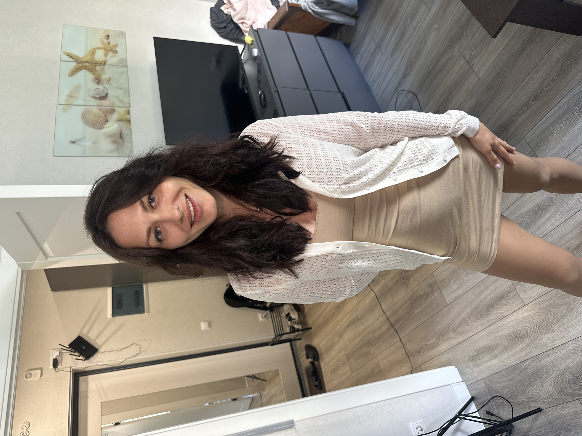 This is a portrait of a young woman with long, dark, wavy hair and light blue eyes. She is smiling and looking at the camera. She is wearing a light beige, form-fitting dress and a white, open-knit cardigan. Her right hand is resting on her knee. The scene appears to be indoors, likely a living room or bedroom, with a wooden floor. In the background, there is a television, a chest of drawers, and wall art depicting starfish and seashells. There is also a router plugged into a wall outlet, with wires connected. The lighting suggests it is daytime, and there are no indications of weather. The overall mood is relaxed and casual. There is no visible text in the image.