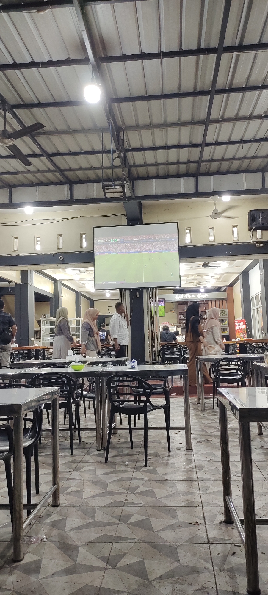 The image depicts an indoor seating area with multiple tables and chairs. A large projection screen hangs from the ceiling, displaying a soccer match with a score of 0-0. Several individuals are present, some standing and observing the screen, while others are seated. The environment appears to be a casual dining or recreational space, possibly a cafe or restaurant, in Keuniree, Indonesia, indicated by a sign that reads "Cafe Maimun Kopi". The ceiling is constructed with corrugated metal panels supported by exposed beams. The floor is tiled with a geometric pattern.