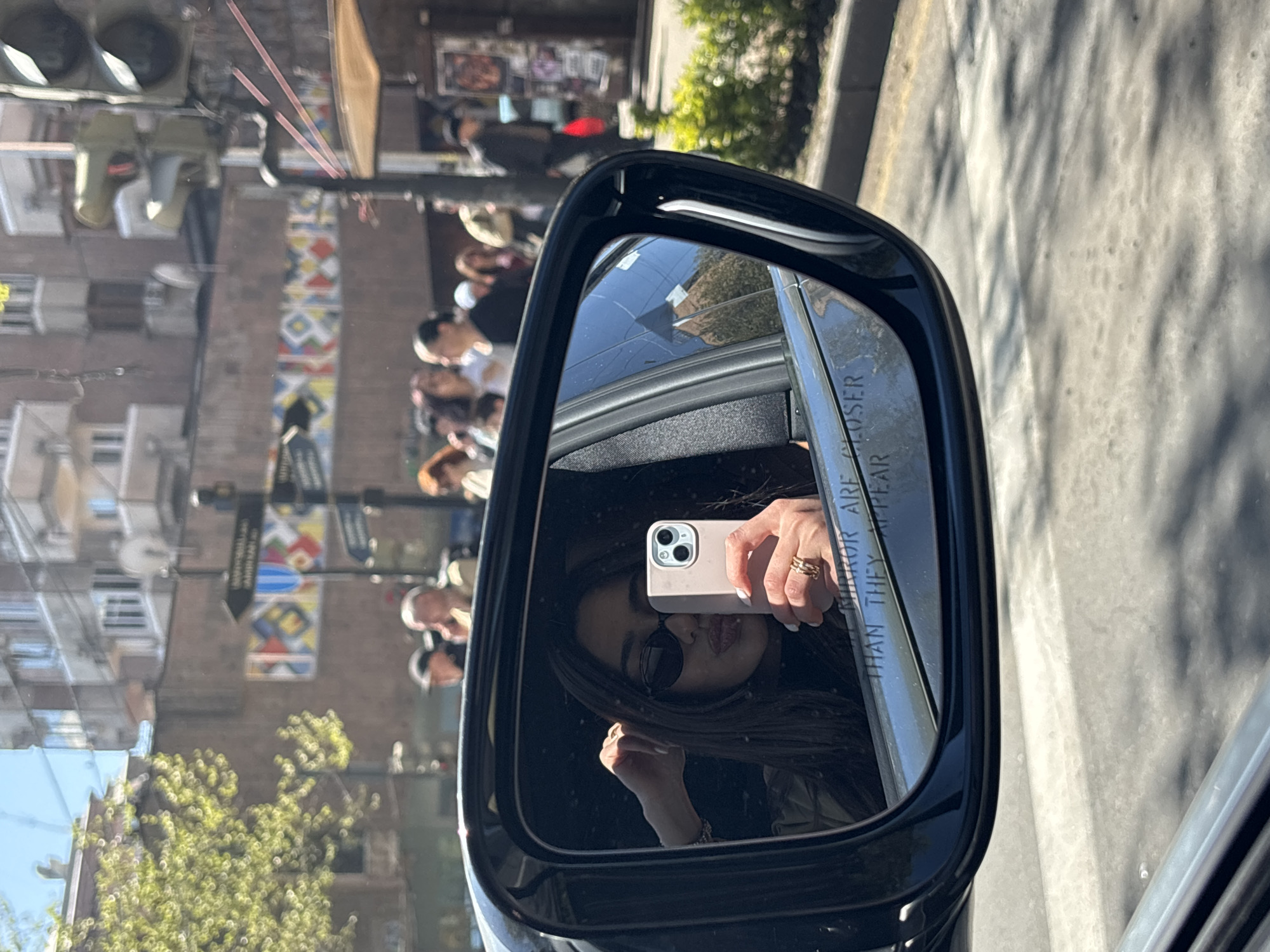 The image depicts a side view mirror of a car, reflecting a woman taking a selfie with her smartphone. She is wearing sunglasses and a ring is visible on her finger. The mirror's reflection shows the exterior environment, including a street scene with buildings, pedestrians, trees, and traffic lights. Based on the architectural style and street banners, the location appears to be in Yerevan, Armenia.