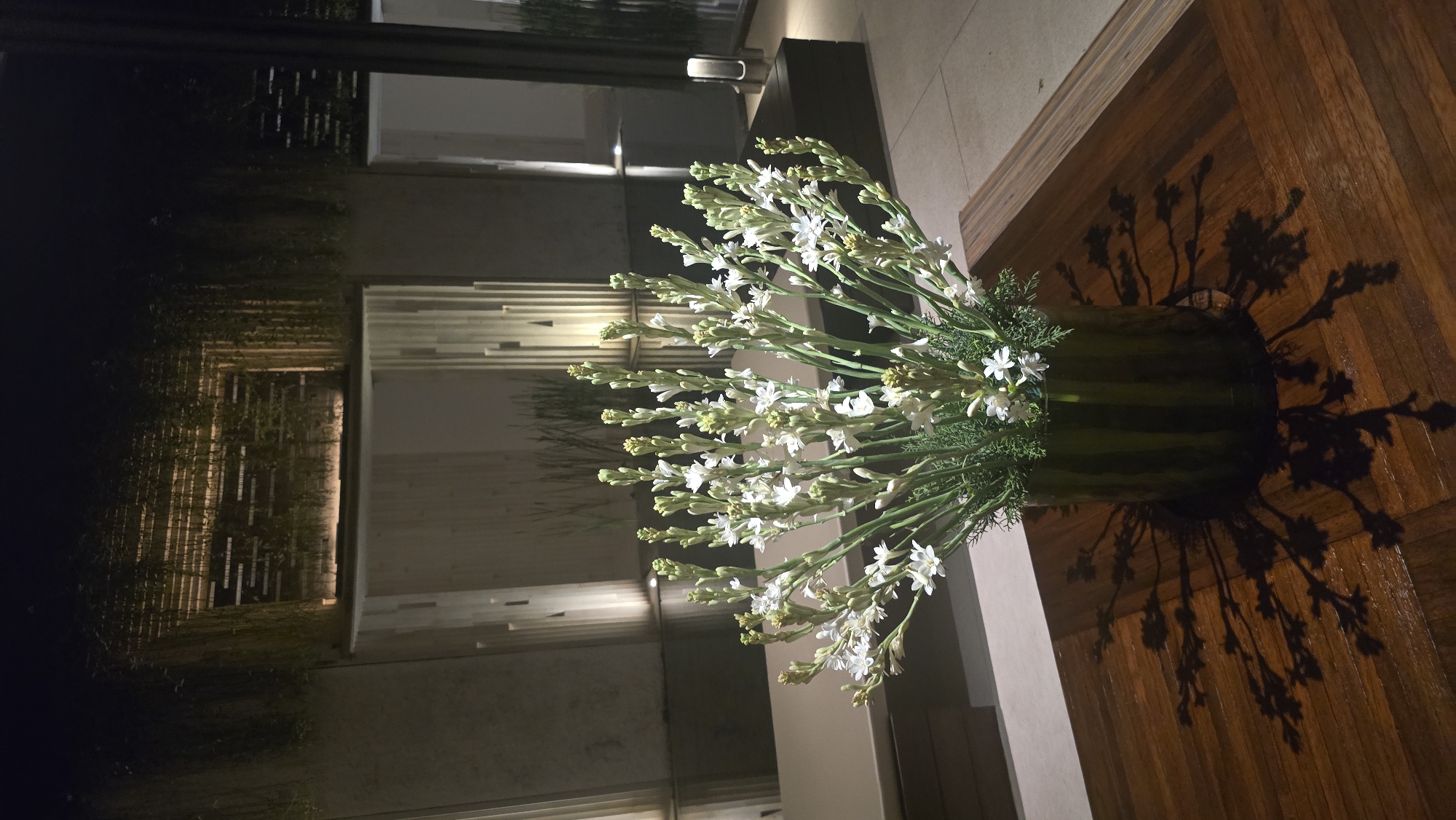 The image shows a floral arrangement with white flowers in a green vase placed on a wooden table. The background includes a building exterior with architectural details such as vertical wooden slats and illuminated sections. The setting is dimly lit, suggesting nighttime. The shadow cast by the floral arrangement is visible on the table. The image was taken in Kuta Selatan, Indonesia.