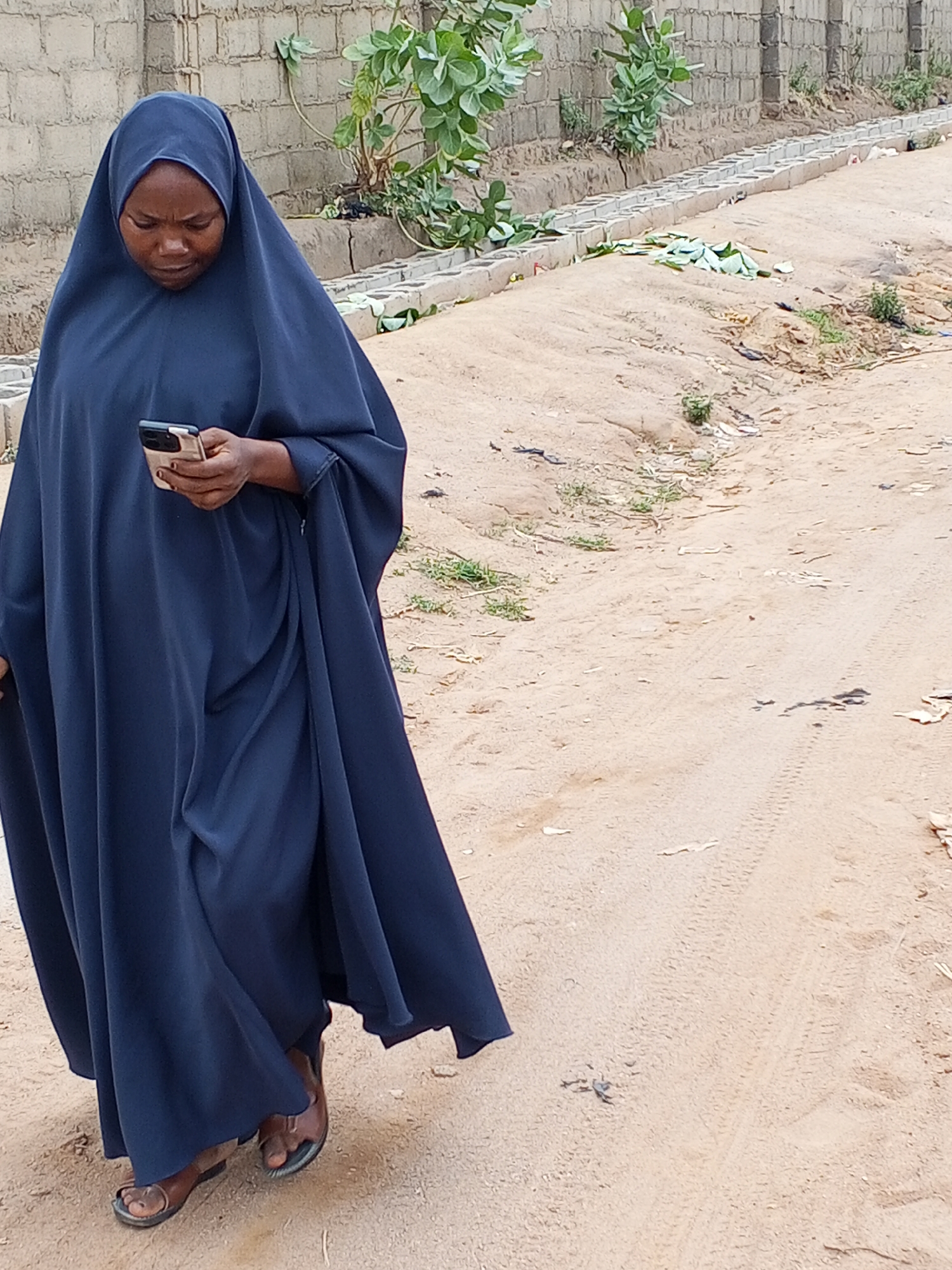 An African woman in Adonkolo, Nigeria, is walking down a dirt road while looking at her smartphone. She is wearing a full-length dark blue garment and brown sandals. The road is sandy, with a few scattered weeds and debris. In the background is a low brick wall with some green plants.