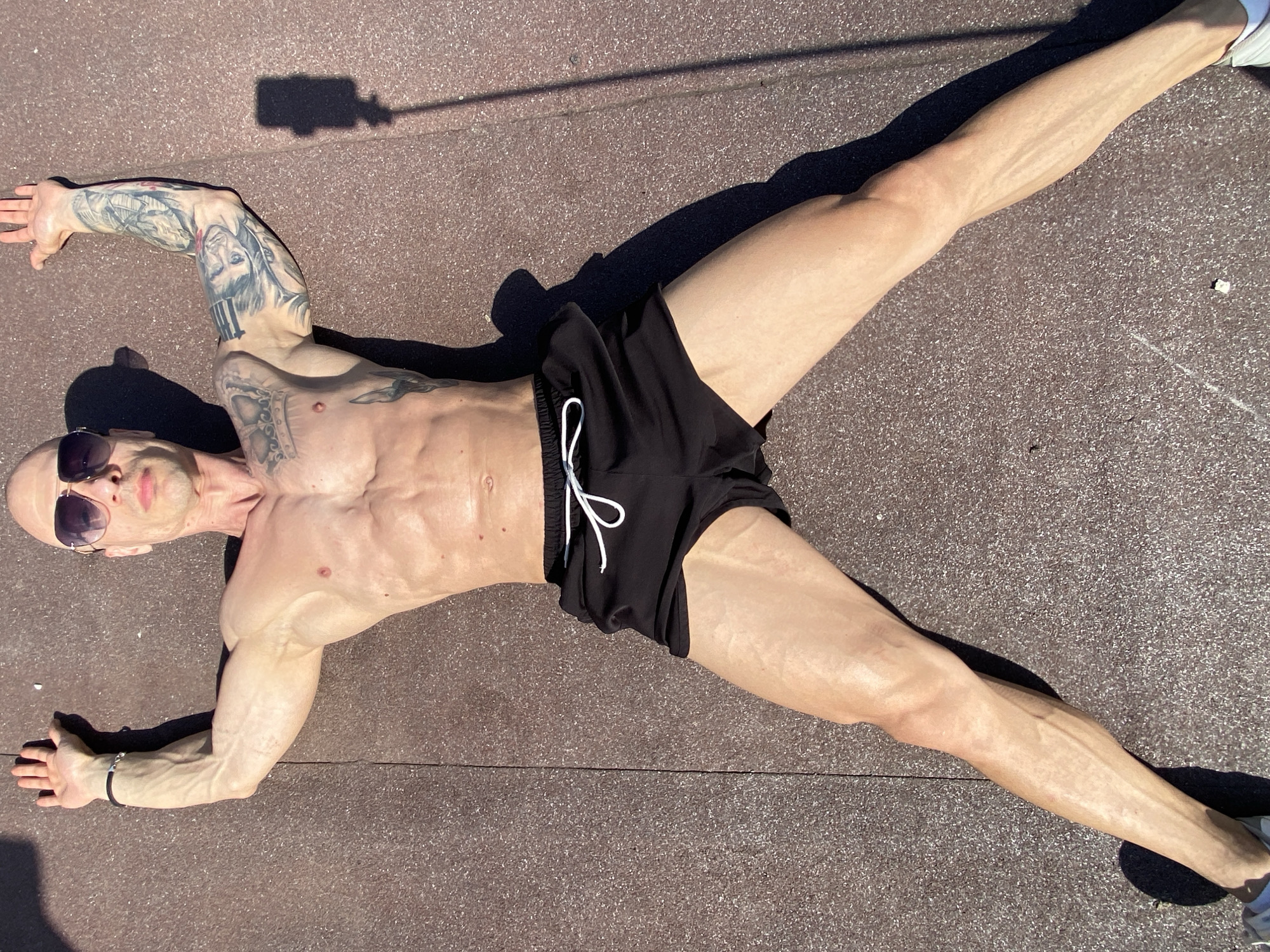 A bald, shirtless man with a lean, muscular physique and pronounced vascularity is lying on his back on a dark reddish-brown, speckled ground surface. He wears dark sunglasses and black athletic shorts with a white drawstring. His arms and legs are spread wide, forming an 'X' shape, with his head slightly turned to his left.
The man has extensive tattoos covering both arms and extending onto his upper torso. His right arm is bent at the elbow, while his left arm is extended outwards. His legs are also spread, with white sneakers visible on his feet. A black bracelet is present on his left wrist.
The outdoor scene is brightly lit by strong sunlight, casting sharp, dark shadows from the man's body and an unseen object in the upper left corner, suggesting a midday or early afternoon setting. The ground appears to be a paved or rubberized surface, potentially a sports court or track. The man's expression is neutral, partially obscured by the sunglasses. No visible text is present in the image. The location is in Pleven, Bulgaria.