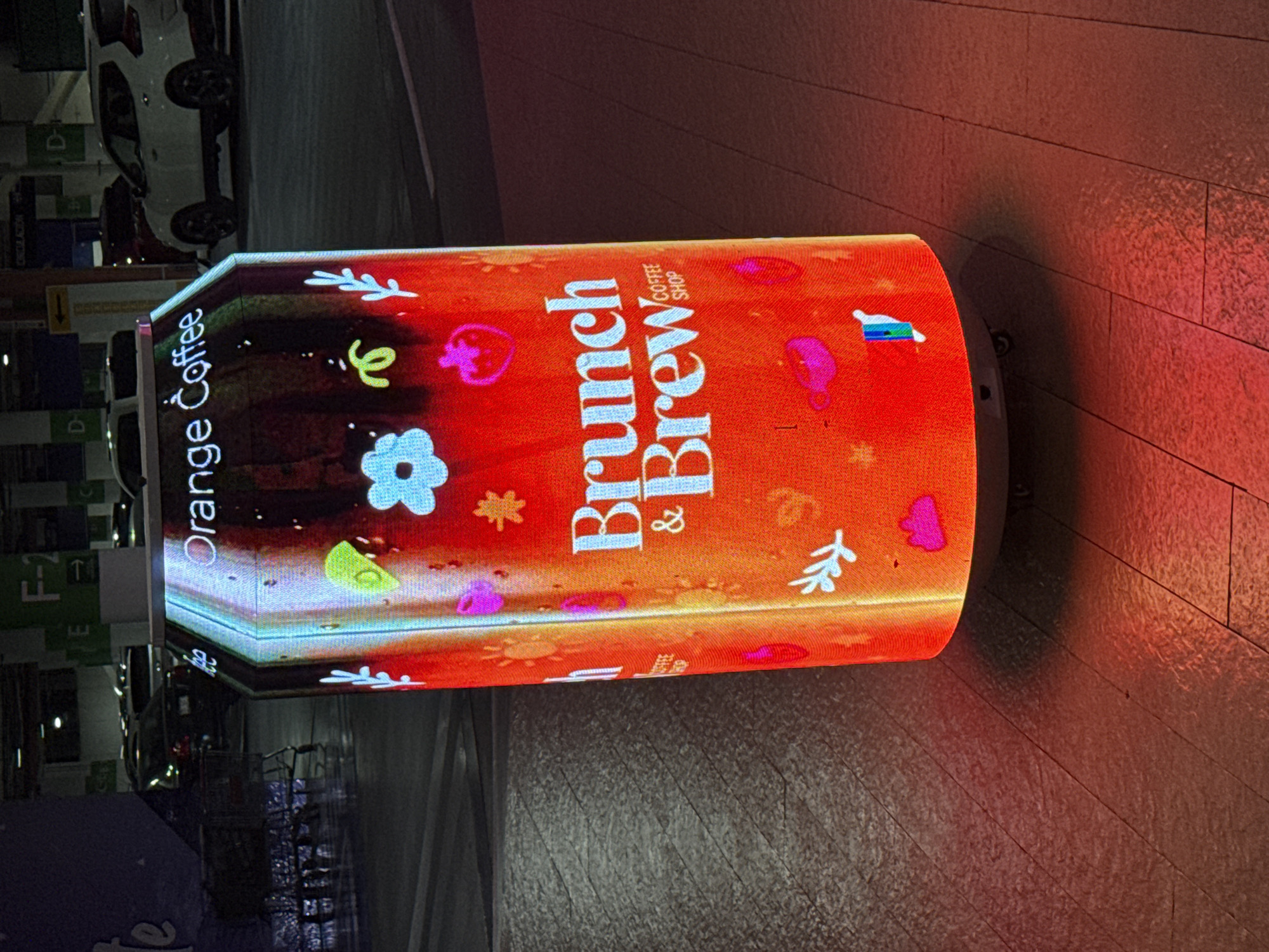 The image shows a cylindrical electronic display with an orange background and white text reading "Brunch & Brew Coffee Shop." The display is decorated with floral and other graphic elements. The display appears to be sitting on a tiled or paved surface. A blurry background suggests an indoor or semi-indoor location, possibly a shopping mall. There is a parking garage with vehicles visible in the background. The location is likely in Puebla, Mexico.