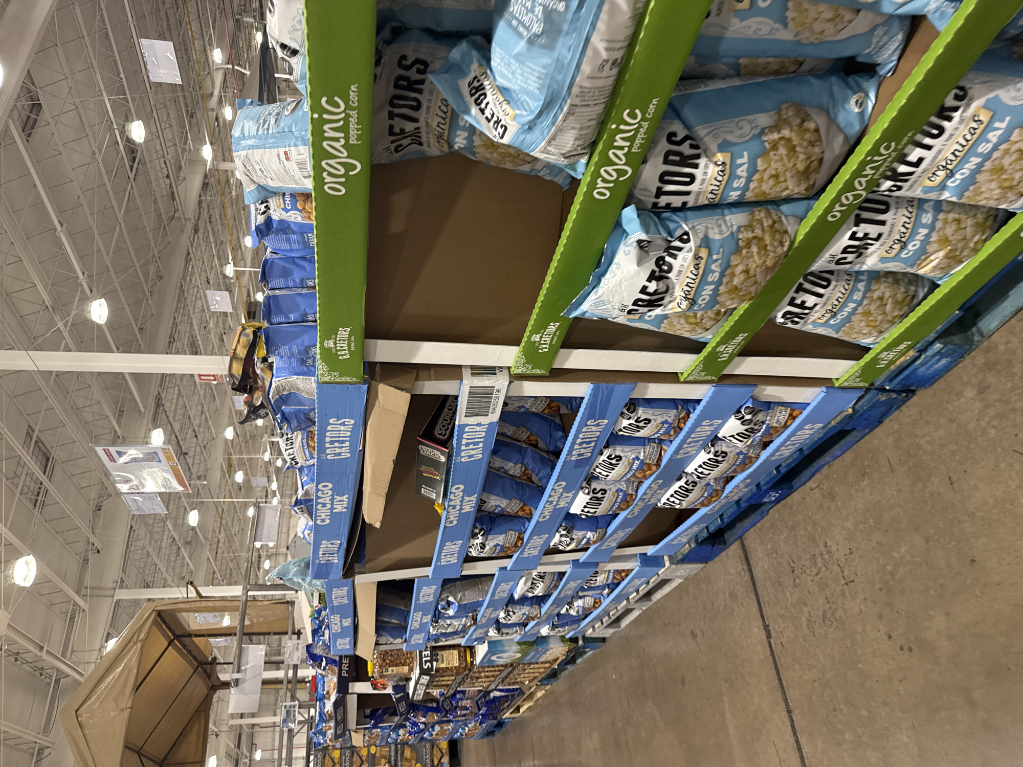 The image shows a shelf in what appears to be a warehouse or large retail store. The shelves are stocked with various products, including several types of popcorn from the brand "G.H. Cretors," such as Chicago Mix and Organic Sea Salt. The packaging includes both English and Spanish text, indicating a possible bilingual market. The environment suggests a busy commercial setting with the aisles filled with different types of snacks and food items. It is unknown if the location is in Puebla, Mexico as there is no indication of this in the image.
