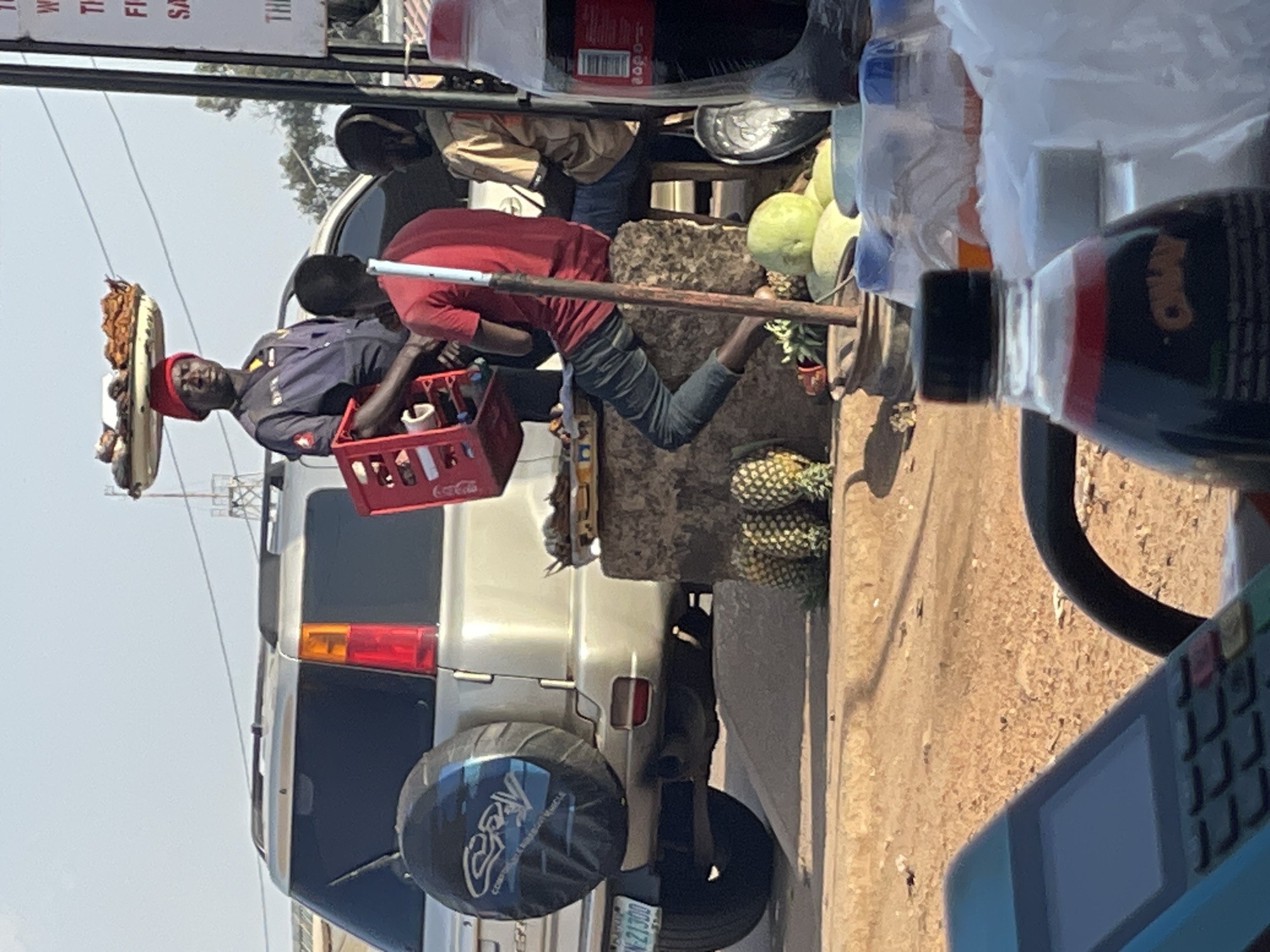 The image depicts an outdoor street scene in Bukuru, Nigeria. A man wearing a police uniform, a red cap, and carrying a tray of food on his head holds a red Coca-Cola crate with bottled drinks while standing near a silver SUV. Another man in a red shirt is leaning over a stone counter, possibly buying pineapples. The counter displays various fruits, including watermelons and pineapples. A third man stands behind the counter. The SUV has a covered spare tire on the back and a visible license plate. In the foreground, part of a point-of-sale (POS) device and bottled drinks are visible.