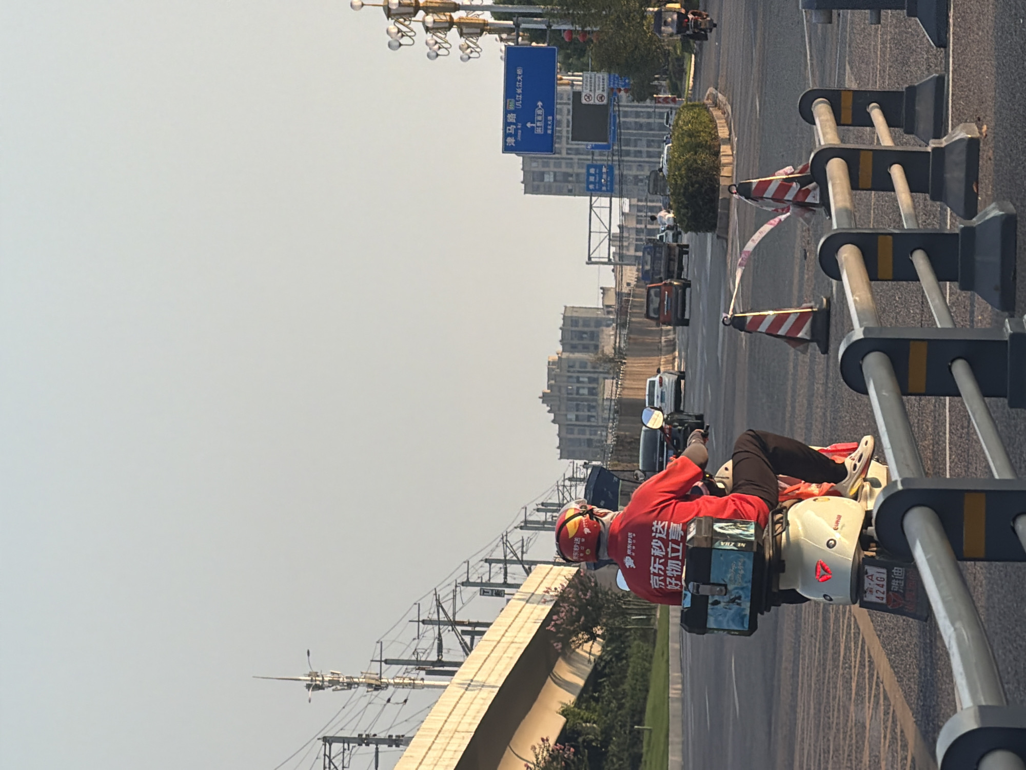 In Jiangjin, China, a delivery person wearing a red shirt with "JD Logistics" branding rides a white electric scooter on a road. The scooter has a blue cargo box on the back, and the delivery person wears a helmet with similar branding. The road has multiple lanes and traffic, including cars and trucks. Traffic cones and barriers are visible, indicating potential road work or a detour. In the background, buildings and elevated train tracks can be seen under a clear sky. A blue road sign indicates directions to "Jinma Road" and other locations. The scene appears to be during daytime, with sunlight casting shadows.