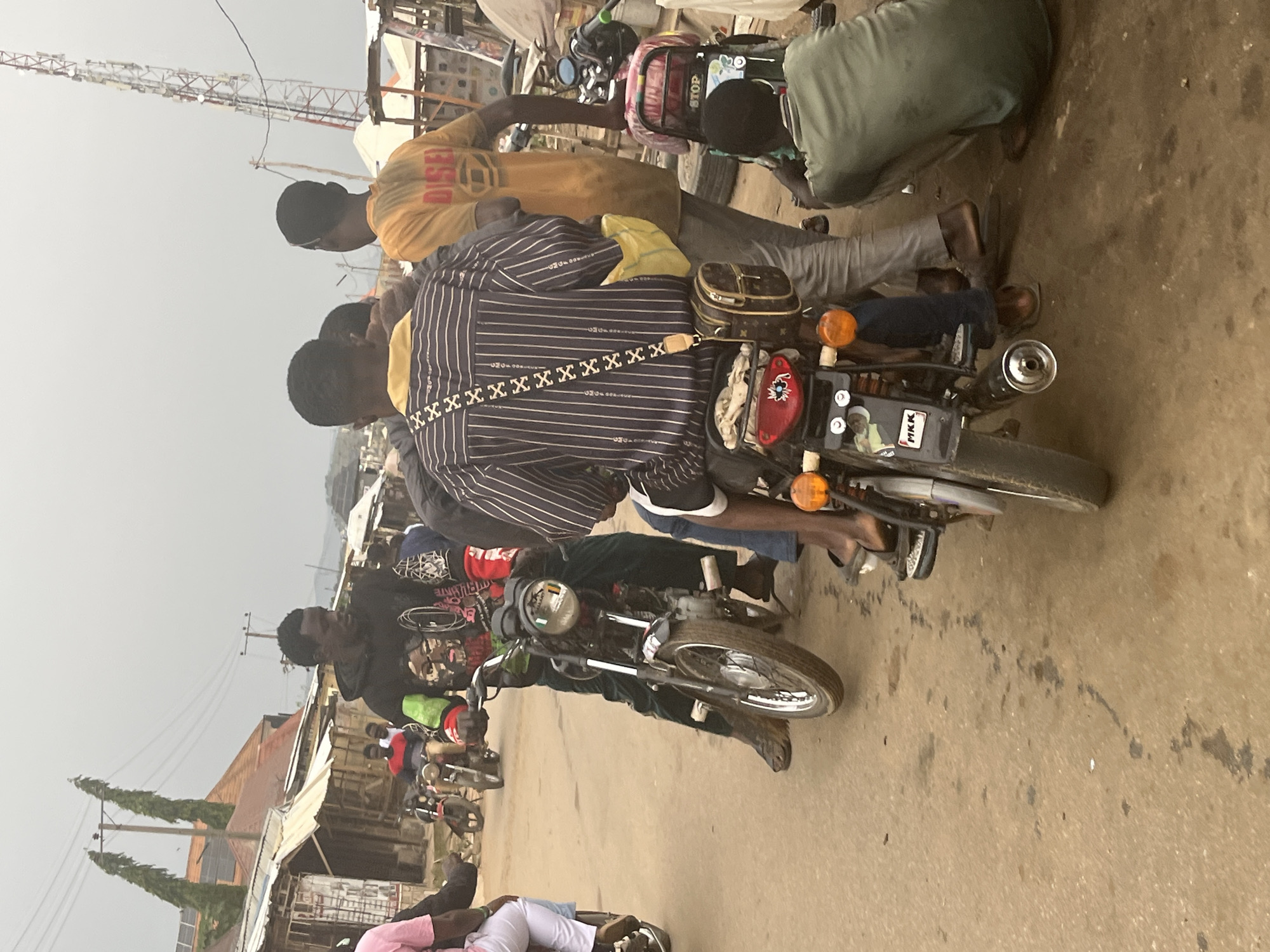 In Abuja, Nigeria, a street scene unfolds with several individuals on and around motorcycles. The focal point is a man in a black hooded sweatshirt adorned with a graphic image, seated on a motorcycle. He is wearing dark pants and brown shoes. To his left, another man in a pink shirt and white pants is partially visible, also on a motorcycle. Behind the man in the black hoodie, a group of individuals are gathered. One man wears a striped shirt and a cross-body bag. Another person is dressed in a yellow t-shirt with "DISEL" printed on it. A person in a green t-shirt is crouched near a motorcycle. The environment appears to be a marketplace or street with modest buildings and structures lining the background. A telecommunications tower is visible in the distance. The ground is paved with asphalt and shows signs of wear and some dark stains. The sky is overcast, contributing to a muted lighting condition. The overall scene depicts a common mode of transportation and daily life in the urban setting.