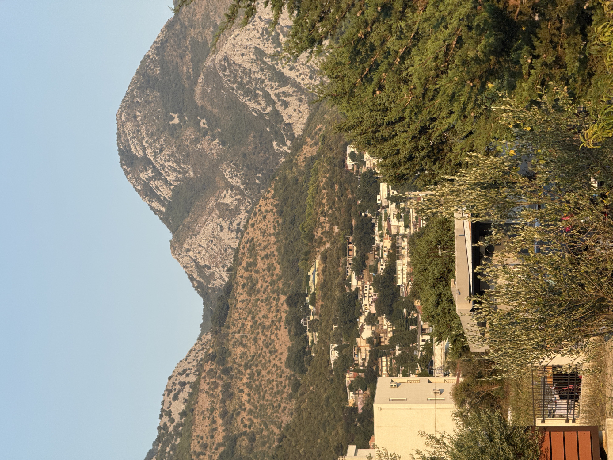 The image depicts a scenic view of Sutomore, Montenegro, with a town nestled on a hillside below rugged mountains. The foreground features lush green foliage, including trees and shrubs, with hints of a fence and part of a building on the left. In the midground, a cluster of white and beige buildings, characteristic of Mediterranean architecture, spreads across the slope. The background is dominated by imposing mountains with rocky outcrops and sparse vegetation, under a clear blue sky. The overall impression is one of a peaceful, sun-drenched coastal town embraced by dramatic natural landscapes.