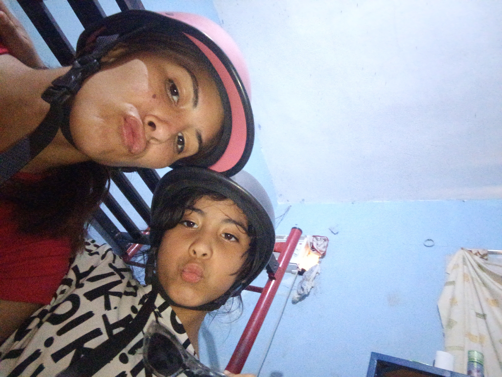 A selfie captured in a building in Juan Griego, Venezuela, shows two individuals wearing helmets. The person in the upper part of the frame has on a pink helmet. The lower individual wears a dark helmet and a white shirt with black geometric designs. A railing is partially visible behind the upper person. In the background, the blue interior includes a ladder-like structure, a patterned cloth, and various objects, including what appears to be cleaning supplies on a shelf. Both subjects are pouting for the photo.