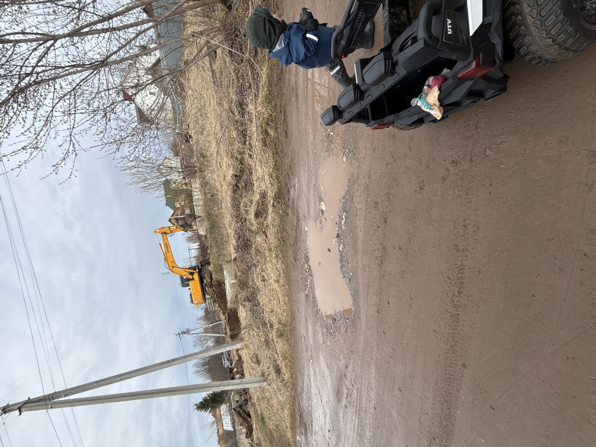 The image depicts a dirt road in the city of Ферма, Russia. A child in a blue jacket and dark hat is standing next to a black UTV. The road is muddy with a large puddle. In the background, there is an excavator, power poles, and vegetation. The sky is overcast.
