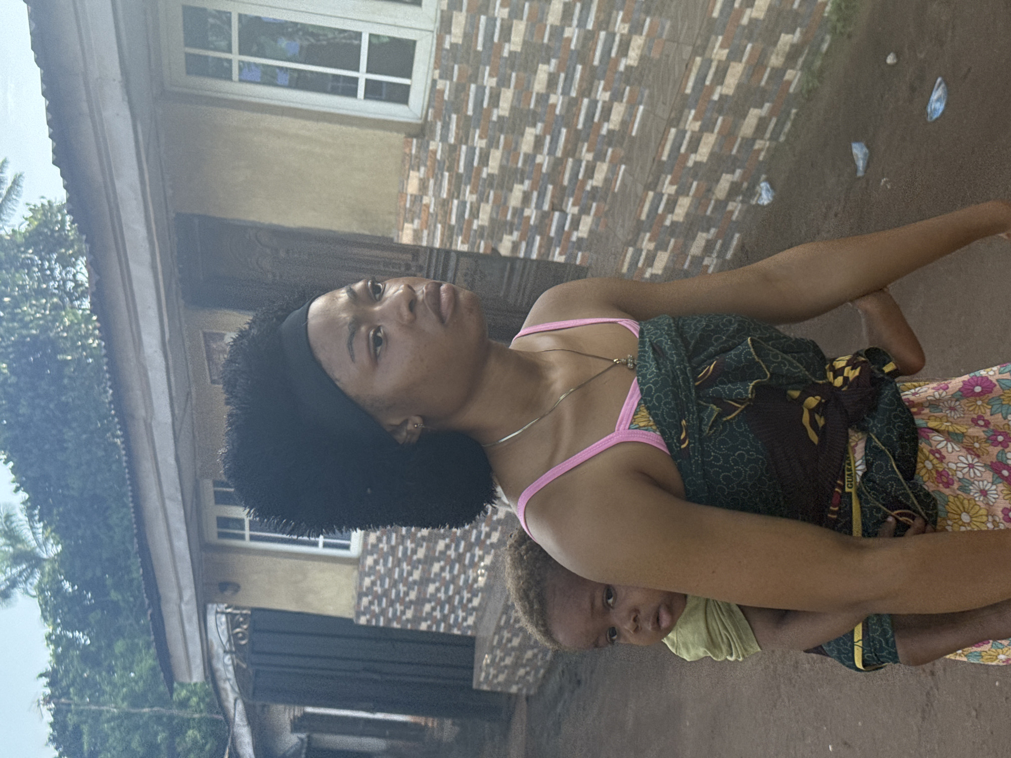 A medium shot depicts a woman carrying a baby outdoors in Aboh, Nigeria. The woman has dark hair and is wearing a pink top with a green and yellow patterned cloth draped around her. She is looking upwards. The baby is blonde, wearing a light green shirt and partially obscured by the cloth. In the background, there is a house with a brown and tan patterned facade. The environment shows greenery and debris on the ground.