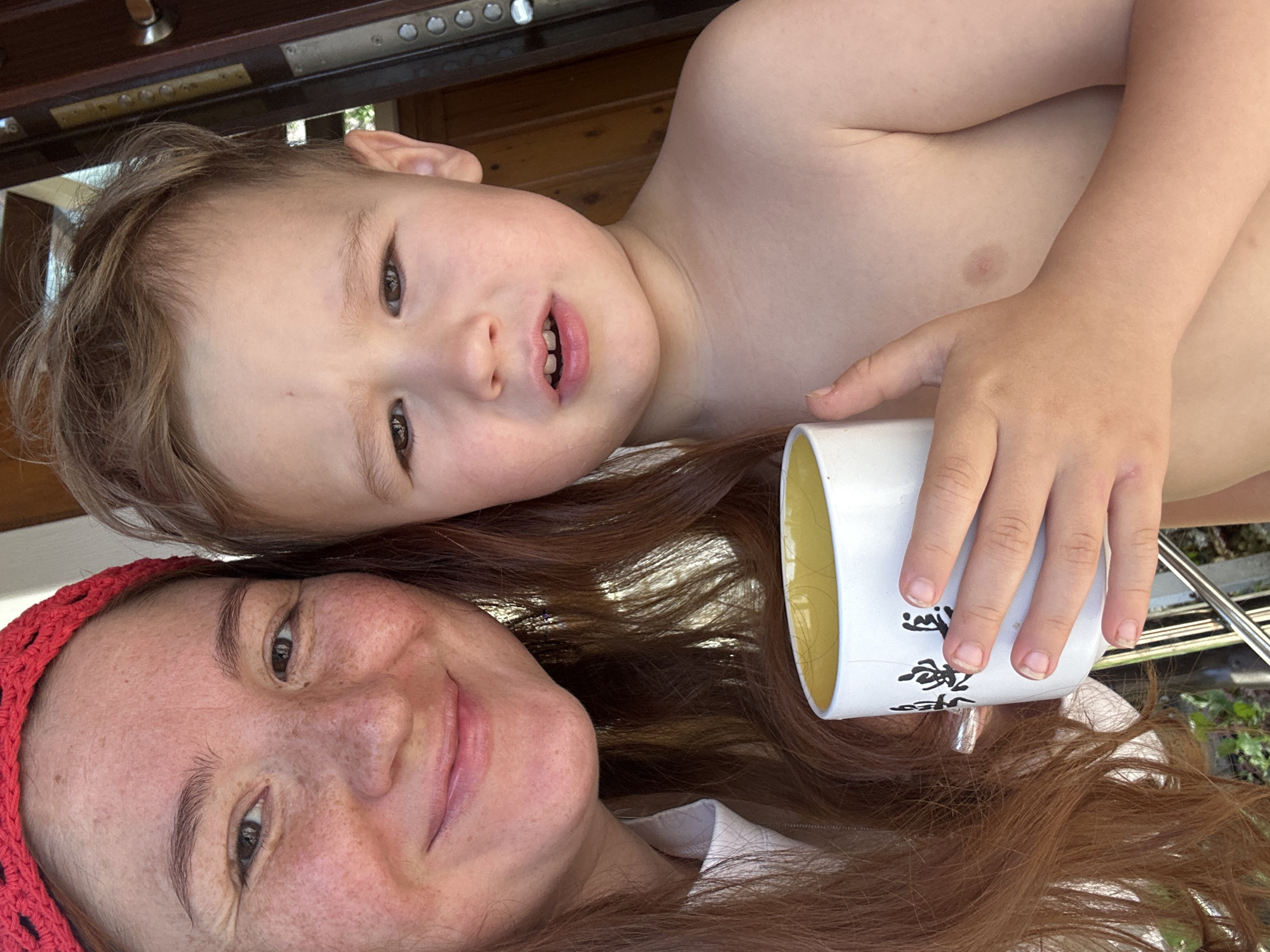 The media file is a selfie taken by a woman and a young child. The woman is positioned at the bottom of the frame, looking towards the camera with a gentle smile. She has reddish-brown hair, some freckles on her face, and is wearing a red crocheted head covering. The child is positioned above her, lying down, with their head turned towards the camera. The child has light brown hair and appears to be looking at something to the side, with their mouth slightly open. The child is shirtless. The woman is holding a white mug with yellow inside. The background appears to be outdoors, with some wooden furniture and greenery visible. The lighting suggests it is daytime, possibly a sunny day. The overall mood is casual and affectionate, likely depicting a moment between a mother and child. There is some Japanese or Chinese text visible on the white mug.