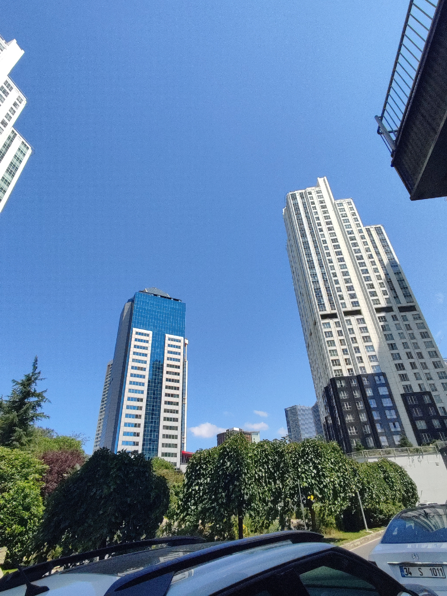This image captures a street-level view looking upwards at a collection of modern high-rise buildings against a clear blue sky. In the foreground, the roofs of two cars are visible. To the left, a white building with multiple windows is partially visible. Further back and to the left, a prominent tall skyscraper with a blue glass facade and white vertical elements stands out. To the right of this blue-clad building, an even taller, mostly white skyscraper with a dark, patterned base dominates the frame. A section of a pedestrian overpass with a railing is visible in the upper right corner. Lush green trees and some shrubbery are positioned in front of the buildings, creating a contrast between nature and urban architecture. The license plate of the white car in the foreground reads "34 S 1011". The scene is likely located in Sarıyer, Turkey, given the visible license plate format which is common in Istanbul.