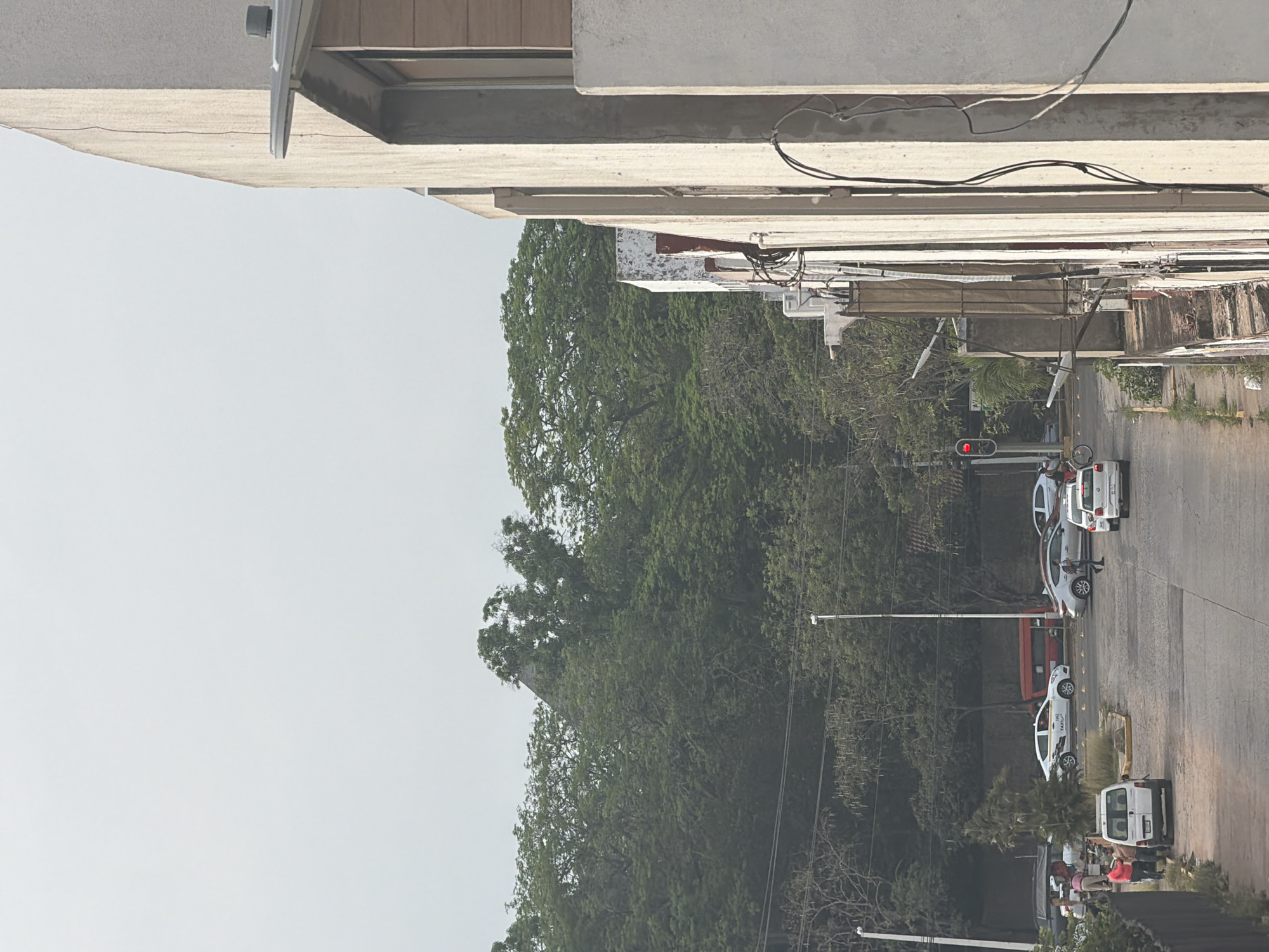 The image depicts a street scene in Villahermosa, Mexico. The view is oriented with the sky on the left, and the road and sidewalk on the right. Dense green trees line the left side of the road. On the right, a line of white cars is parked on the street, including at least one taxi. A red traffic light is visible further down the road. In the foreground, a building overhangs the scene. The environment appears to be urban with a mix of residential or commercial buildings and natural green spaces. There are people on the sidewalk near a white delivery truck.