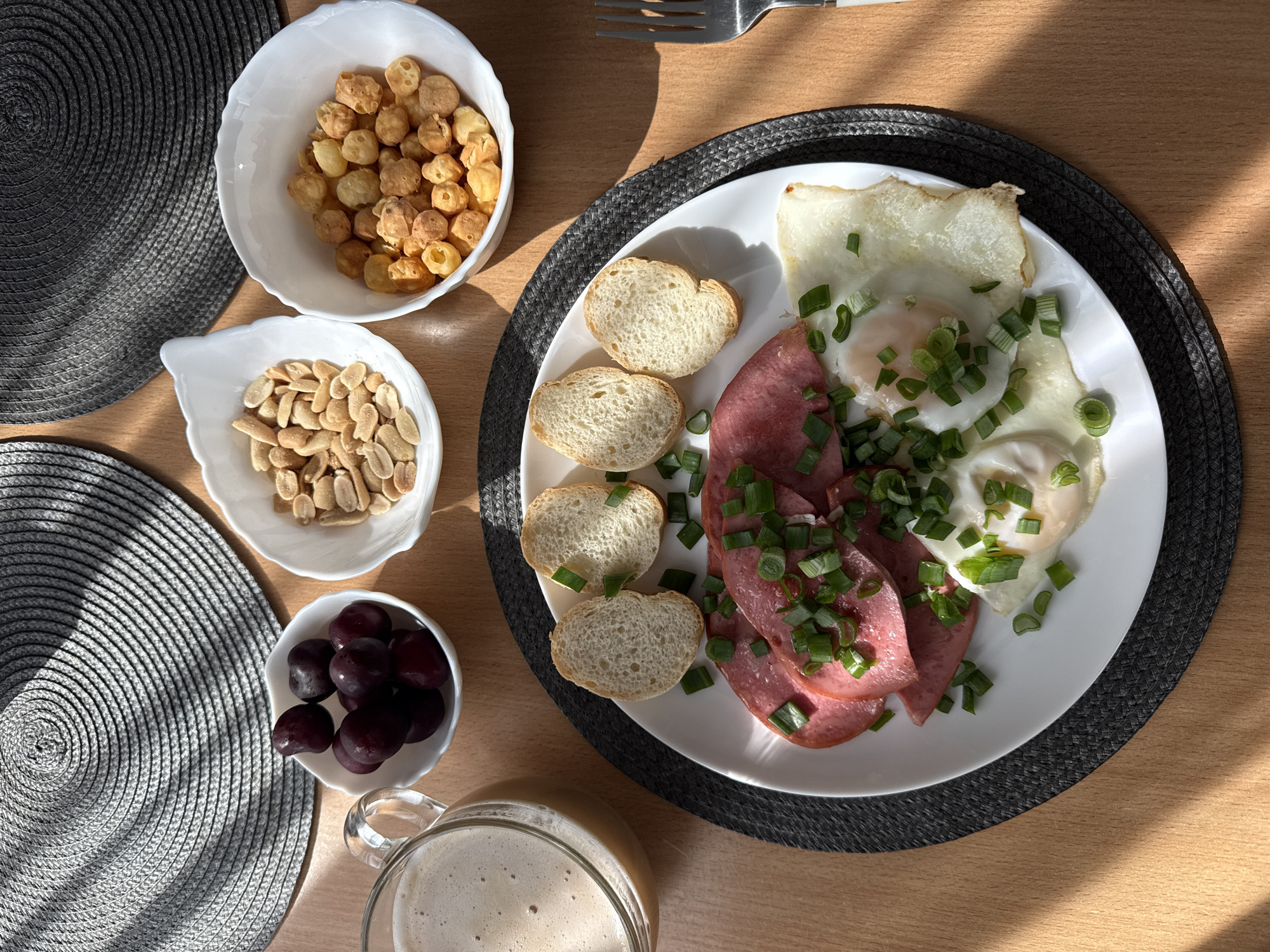 A top-down shot captures a breakfast spread on a wooden table. In the foreground, a white plate holds a meal consisting of three fried eggs, sliced cured meat garnished with chopped green onions, and four small slices of bread. To the left of the plate is a clear glass mug filled with a frothy, light-brown beverage.
The meal is set on a round, dark grey placemat. Behind the plate and mug, to the left and right, are two more round placemats with a textured, woven design.
In the background, arranged in a line above the plate, are three small white bowls. The leftmost bowl contains dark red cherries. The middle bowl is filled with roasted peanuts. The rightmost bowl contains small, puffy, golden-brown snacks. Sunlight casts shadows from the woven placemats onto the table surface. A silver fork rests to the right of the plate. The scene is set indoors, with no identifiable people present and no specific city or location information available.
