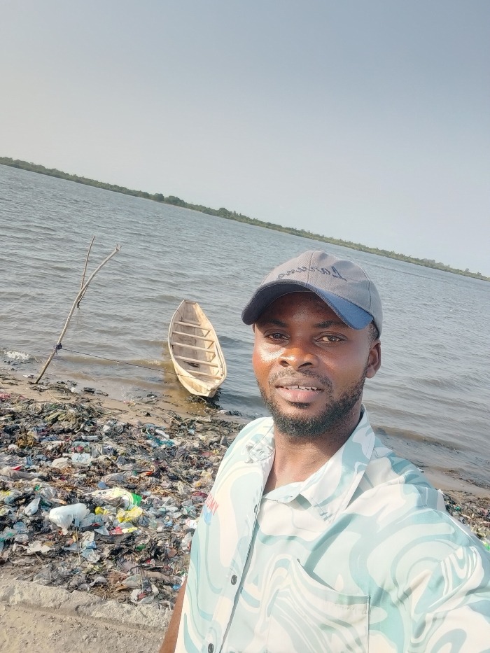 A man is taking a selfie outdoors by a body of water on what appears to be a bright, clear day. He is positioned in the foreground, facing the camera, with a slight smile on his face. He has dark skin, a short beard, and is wearing a light-colored, short-sleeved collared shirt with a wavy, abstract pattern in shades of light blue and white. He is also wearing a grey baseball cap with a blue brim. On the cap, text is partially visible, appearing to be "Ommuso" or similar.
Behind the man, to his left, is a small, light-colored wooden dugout canoe or boat, partially in the water and partially on the muddy/sandy shore. A thin, branched stick or pole is stuck into the ground near the water's edge, slightly to the left of the boat.
The shoreline in the lower left and central-left of the image is heavily littered with a large accumulation of various types of trash, including plastic bags, bottles, and other debris, creating a significant environmental blight. The water beyond the shore is a wide expanse of a lake or river, with gentle ripples visible on its surface. In the far background, across the water, a distant landmass or shoreline with green vegetation (trees or bushes) is visible under a clear, bright sky. The overall impression is of a natural landscape significantly impacted by pollution.