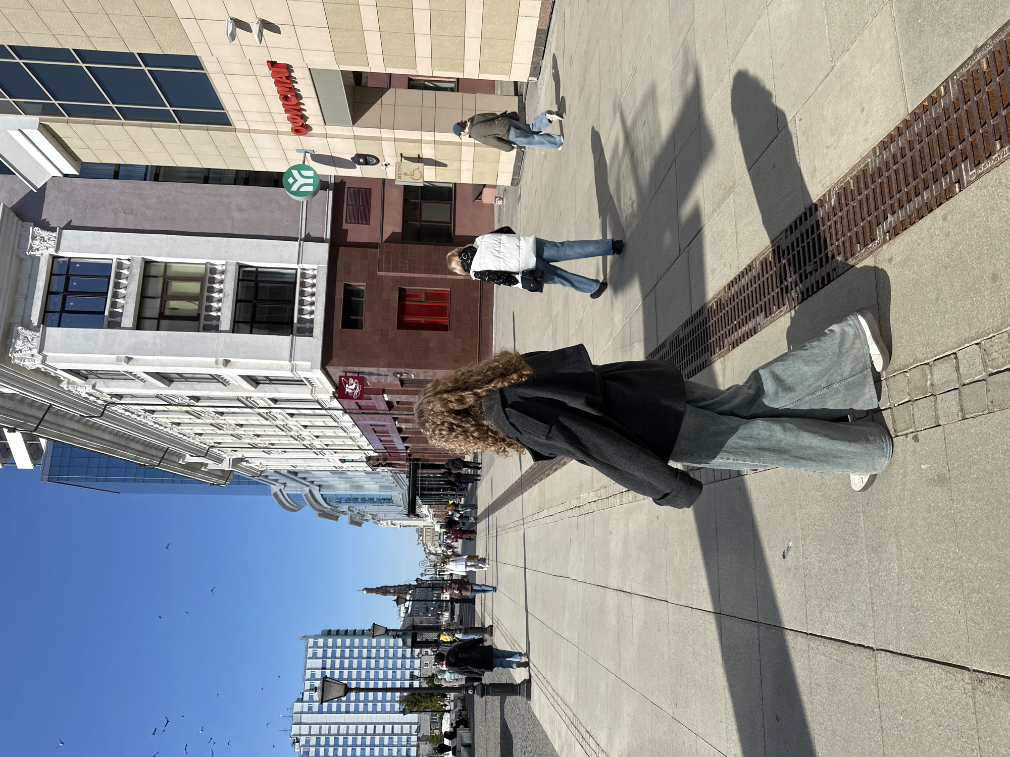The image depicts a bright, sunny day on Bauman Street in Kazan, Russia, a pedestrian thoroughfare. The scene is dominated by a person in the immediate foreground, observed from a rear-quarter angle, walking from right to left across light gray concrete pavers. This individual has long, curly, reddish-brown hair and is dressed in a dark grey short jacket, light blue wide-leg jeans, and white athletic shoes with brown soles. Their shadow is distinctly cast towards the right, falling partly on a long, rusty metal drainage grate embedded in the pavement.
Further down the street, on the right side, another person walks away from the camera, wearing a white puffer vest over a black and white patterned hoodie, light blue wide-leg jeans, and dark shoes, carrying a black shoulder bag. Behind them, a third individual, clad in a green jacket, blue baseball cap, and blue jeans, also walks away from the camera, carrying a dark shoulder bag.
On the left side of the street, multiple pedestrians are visible in the midground and background, walking in various directions. Black, ornate street lamps line the left side of the pedestrian path. In the far distance, centered along the street's perspective, the dark, spire-topped Bogoyavlensky bell tower is clearly visible.
Architectural details define the street's edges. On the right, a building with beige paneling features large rectangular windows and a prominent red sign displaying "ОФИСМАТ" in Cyrillic, along with a green circular logo. An accessibility sign with a wheelchair symbol and "МЕТРО" is affixed to a column on this building. Adjacent to it, a building with a white facade showcases classical architectural elements, including columns and balustrades, with a reddish-brown base and a red sign featuring a stylized white logo. Further into the background, modern glass and steel structures rise. On the left, a tall building with a grid pattern of light panels and dark windows stands against the clear blue sky, where several birds are in flight. The sun, casting long shadows, indicates a low angle, consistent with morning or late afternoon.