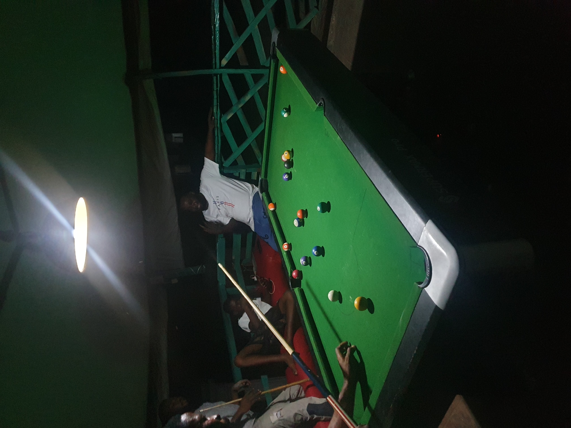 The image depicts a group of individuals engaged in a game of pool at night, likely at an outdoor establishment in Lokoja, Nigeria. A billiard table with green felt is centrally positioned, with multiple colored balls scattered across its surface. Several people are visible, some actively participating in the game, while others are observing or relaxing. One person, wearing a white t-shirt, is leaning on a railing and appears to be gesturing. Another individual is holding a pool cue, poised to take a shot. The setting appears to be covered by a dark canopy, with a bright overhead light illuminating the pool table. The surrounding environment is dimly lit, suggesting a casual, recreational atmosphere.