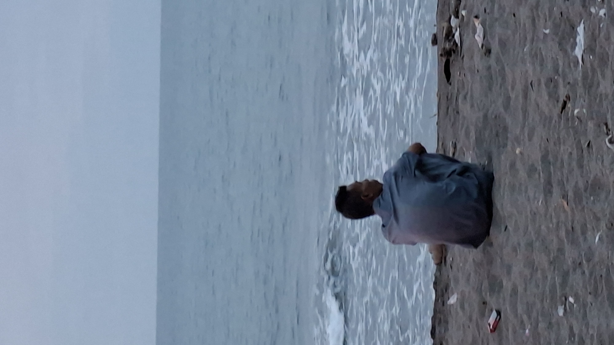 A lone individual, viewed from behind, sits cross-legged on a sandy beach facing the ocean. The person is wearing a light blue shirt. The water appears calm with gentle waves. The beach is littered with various debris, including plastic and what appears to be cigarette packs. The sky is overcast and a dull gray, suggesting it is either early morning or late evening. The location is identified as the Lhokseumawe City beach in Indonesia.