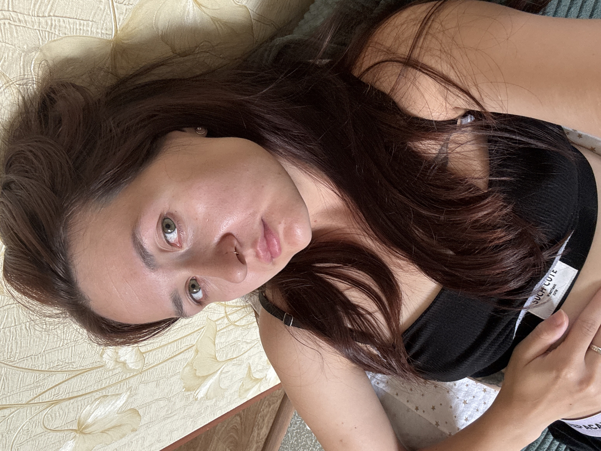 An overhead, close-up shot shows a young woman lying down, looking upwards at the camera. She has dark brown hair and light brown eyes. Her hair is loose and falls around her face and shoulders. She is wearing a black tank top with a white tag that reads "SUCH CUTE". Her left hand is resting on her stomach, and she is wearing a ring on her left index finger. She has a nose ring. The background is a textured, light-colored wall with a floral pattern. The image does not provide any visual cues to determine her location in Naberezhnye Chelny, Russia.