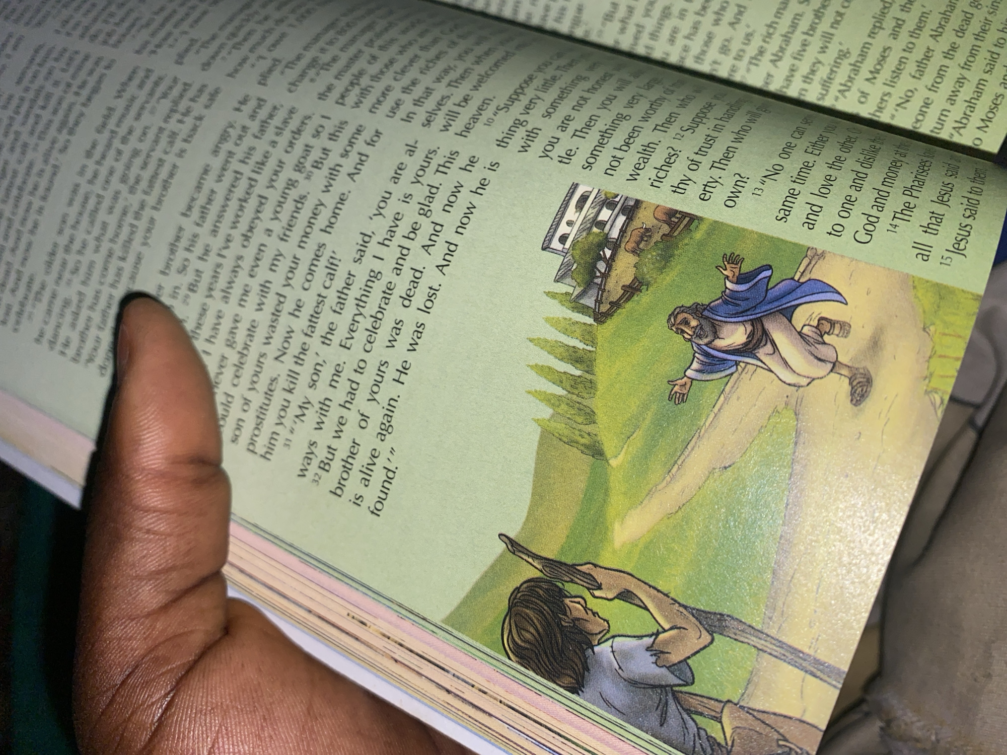 The image shows an open book with a Bible illustration on the left page and text on both pages. A person's hand is holding the top of the book, obscuring some of the text. The illustration depicts a pastoral scene with two figures and a building. On the left, a young man with dark hair, wearing a light-colored tunic, stands facing right, holding a wooden staff. To the right, an older man with a beard and wearing a blue robe and white tunic runs toward the left, with his arms outstretched. In the background, a white building with arched windows and a red-tiled roof is visible, surrounded by a fence and some animals resembling cows. Trees are scattered across the landscape, which includes a dirt path. The right page is filled with text, organized in columns. The book is open to a page with a green border. The lighting suggests an indoor environment. The image does not contain any specific information or visual cues that would allow for the identification of the location as Jauro Yinu, Nigeria.