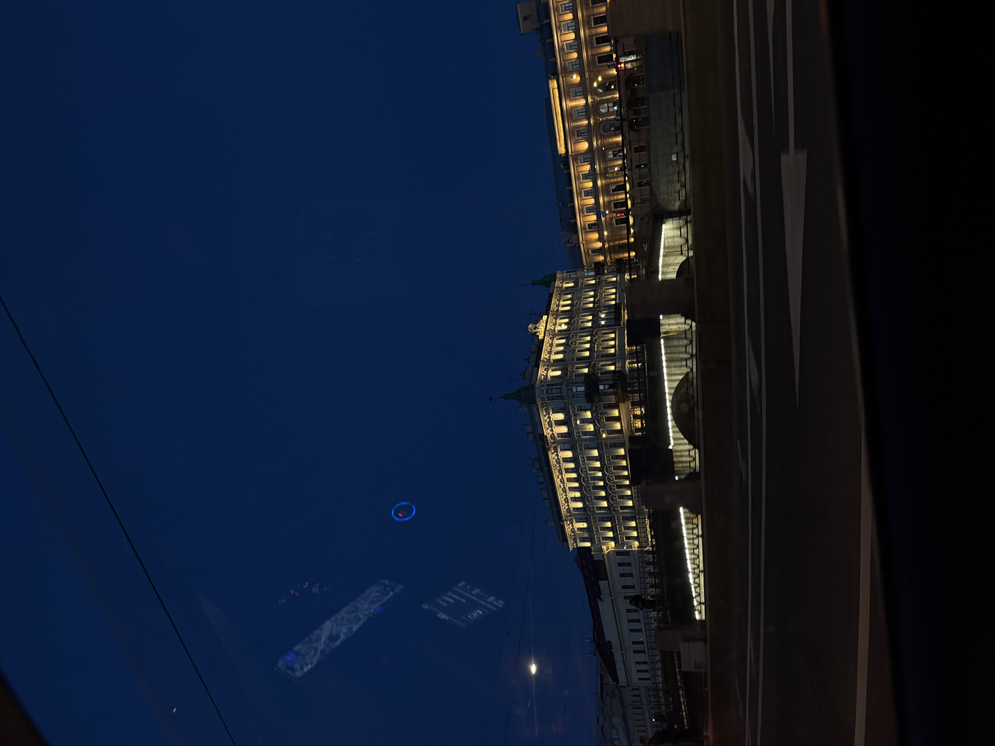 The image shows a street view in Saint Petersburg, Russia, at dusk. The sky is a deep blue, transitioning from twilight to night. In the foreground, a road with white directional arrows is visible, suggesting a driving perspective. Beyond the road, a canal or river is present, with a bridge spanning across it. Along the waterfront, ornate buildings with illuminated windows are seen, typical of Saint Petersburg's architectural style. The buildings are lit up, highlighting their classical facades. Reflections are visible on what appears to be a window pane, showing a blurred image of interior lights and possibly some text. One prominent reflection is a bright blue circular light. The overall scene depicts a well-lit urban environment at night, with the distinctive architecture of Saint Petersburg as the backdrop.