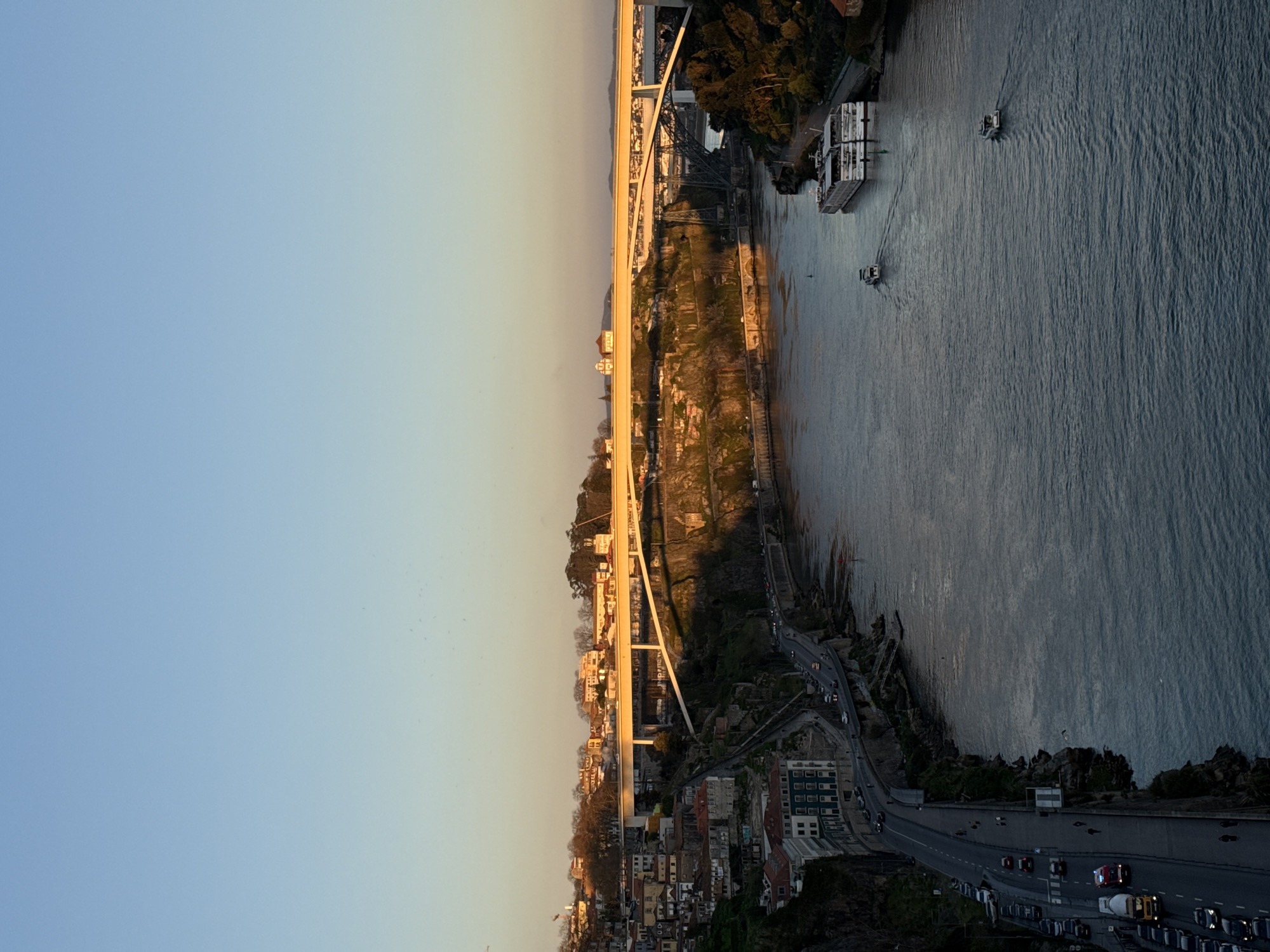 This image captures an elevated, panoramic view of the Douro River in Porto, Portugal, during late afternoon or early evening, characterized by warm, low-angle sunlight.
The sky occupies the upper third of the frame, clear with a gradient from light blue at the zenith to a pale yellowish hue near the horizon, indicative of sunset.
The Douro River flows prominently through the middle ground, its dark blue-grey surface reflecting the sky, with visible ripples and wakes from moving vessels. On the river, one large, white multi-deck passenger cruise ship is docked along the right (north) bank. Two smaller, white motorboats are actively traversing the river upstream, leaving distinct wakes behind them.
The left (south) bank, part of Porto, features a steep, rocky cliff face densely covered with vegetation and numerous multi-story residential buildings with red-tiled roofs, terraced up the hillside. A curved, paved road runs along the base of this cliff, parallel to the river. Numerous passenger cars are visible on this road, some parked and others in motion, heading left (west). A yellow cement mixer truck and an emergency vehicle with blue flashing lights are also visible on the road, traveling in the same direction. Several indistinct figures appear as pedestrians on the sidewalk adjacent to the road.
Three bridges span the river in the mid-ground and background. The most prominent is the Ponte do Infante, a modern white concrete arch bridge, illuminated golden by the setting sun, carrying vehicular traffic. Behind it, further downriver, is the Ponte de São João, a darker metal truss railway bridge, partially obscured by the Ponte do Infante. In the far distance, another large arch bridge, the Ponte da Arrábida, is faintly visible. The right bank primarily shows parts of Vila Nova de Gaia, with scattered buildings and trees illuminated by the setting sun.